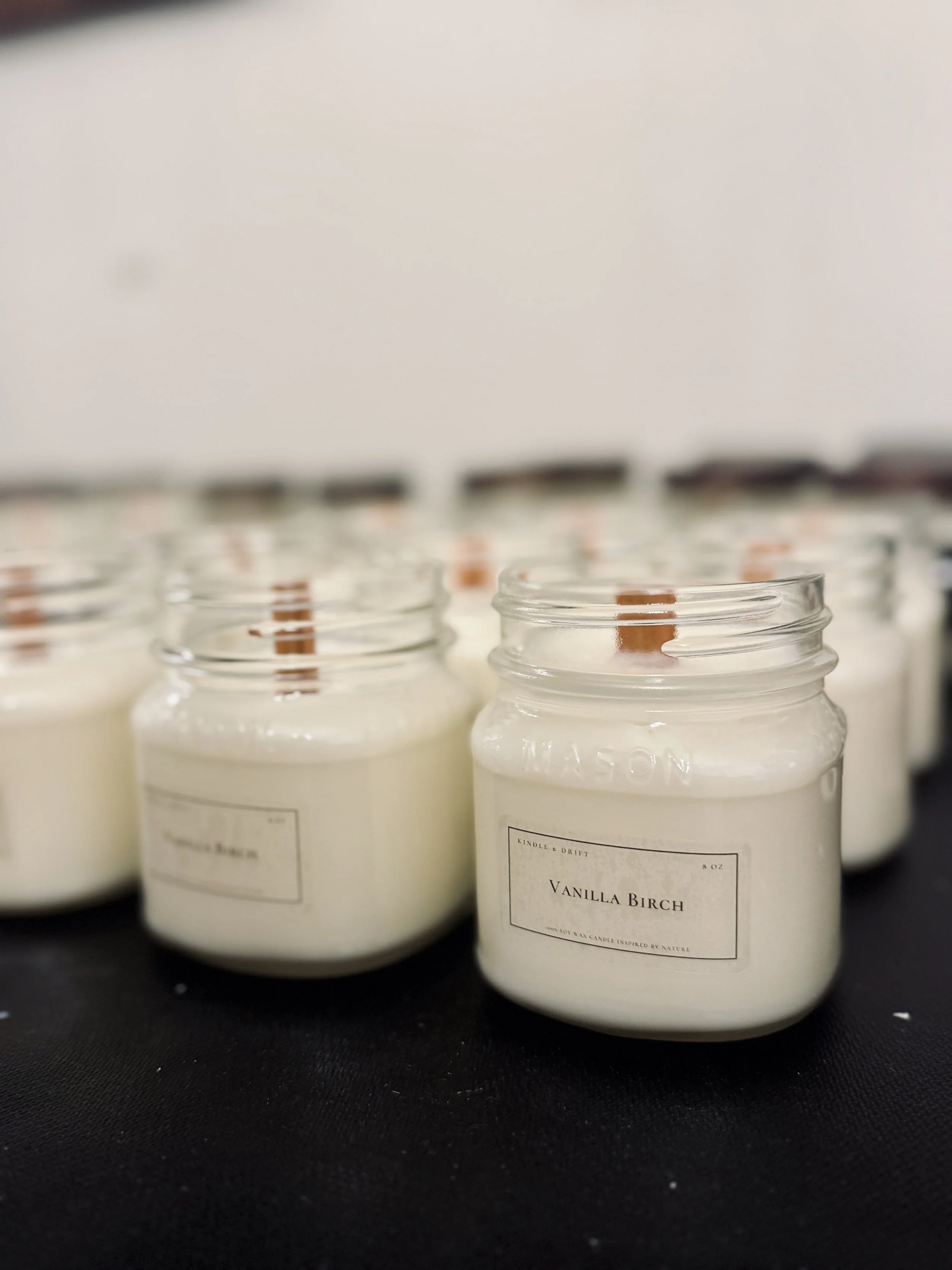 Several white vanilla scented candles in glass jars with wooden wicks, arranged on a black surface.