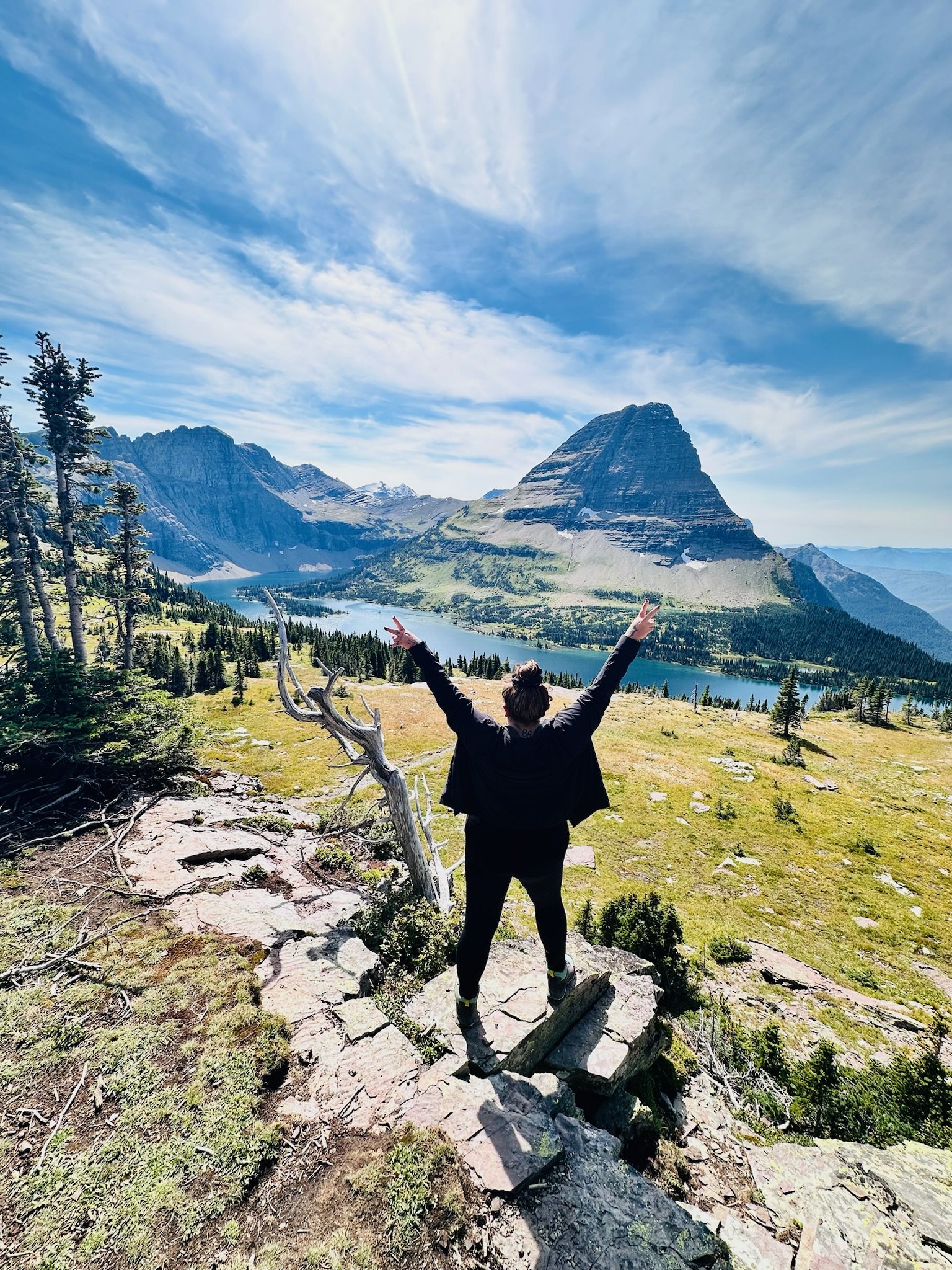 Person standing on rocky trail with arms raised, overlooking a mountain lake with mountain peaks in the distance under a partly cloudy sky.