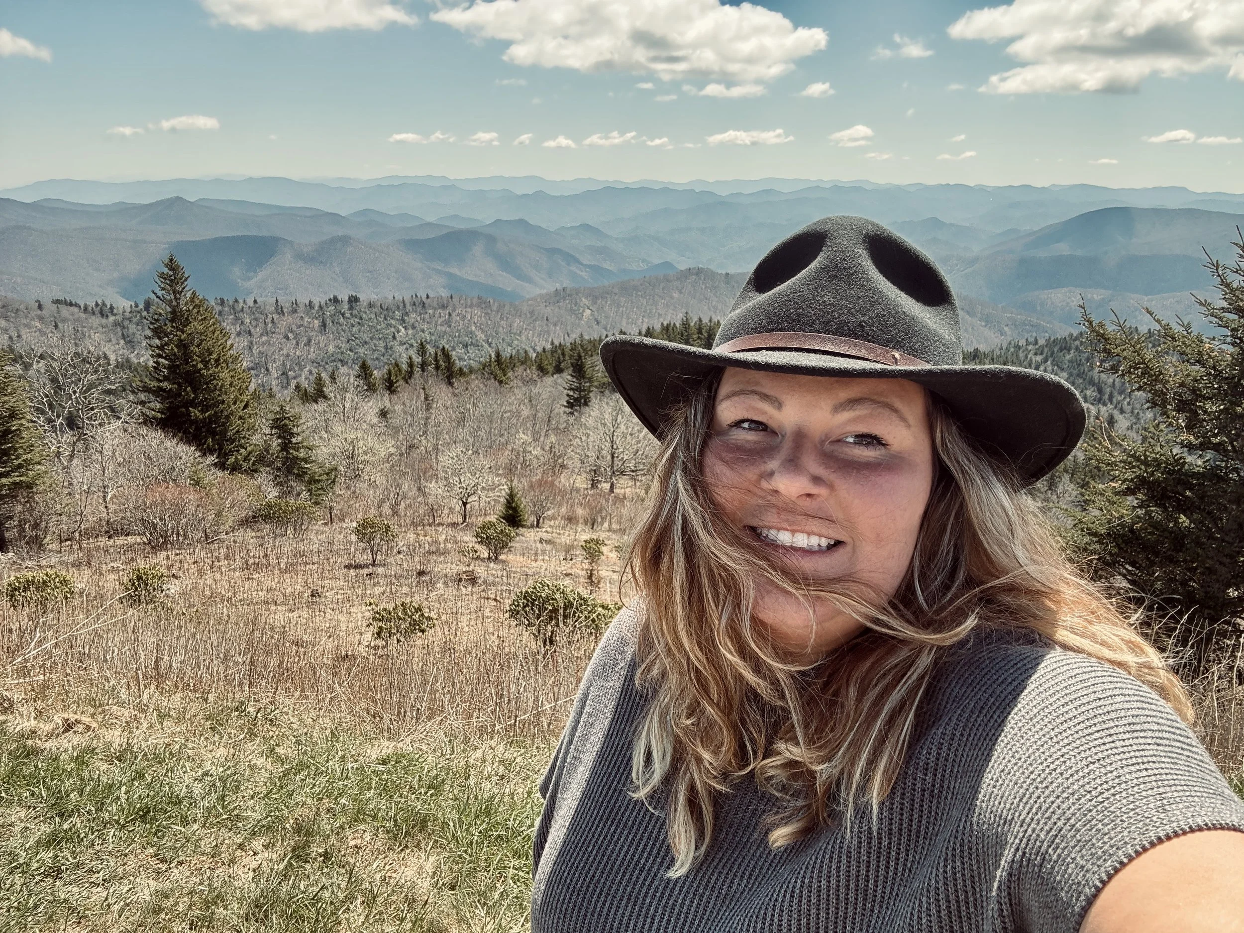 A woman smiling and taking a selfie outdoors with a mountain range and trees in the background. She is wearing a wide-brimmed hat and a short-sleeve shirt.