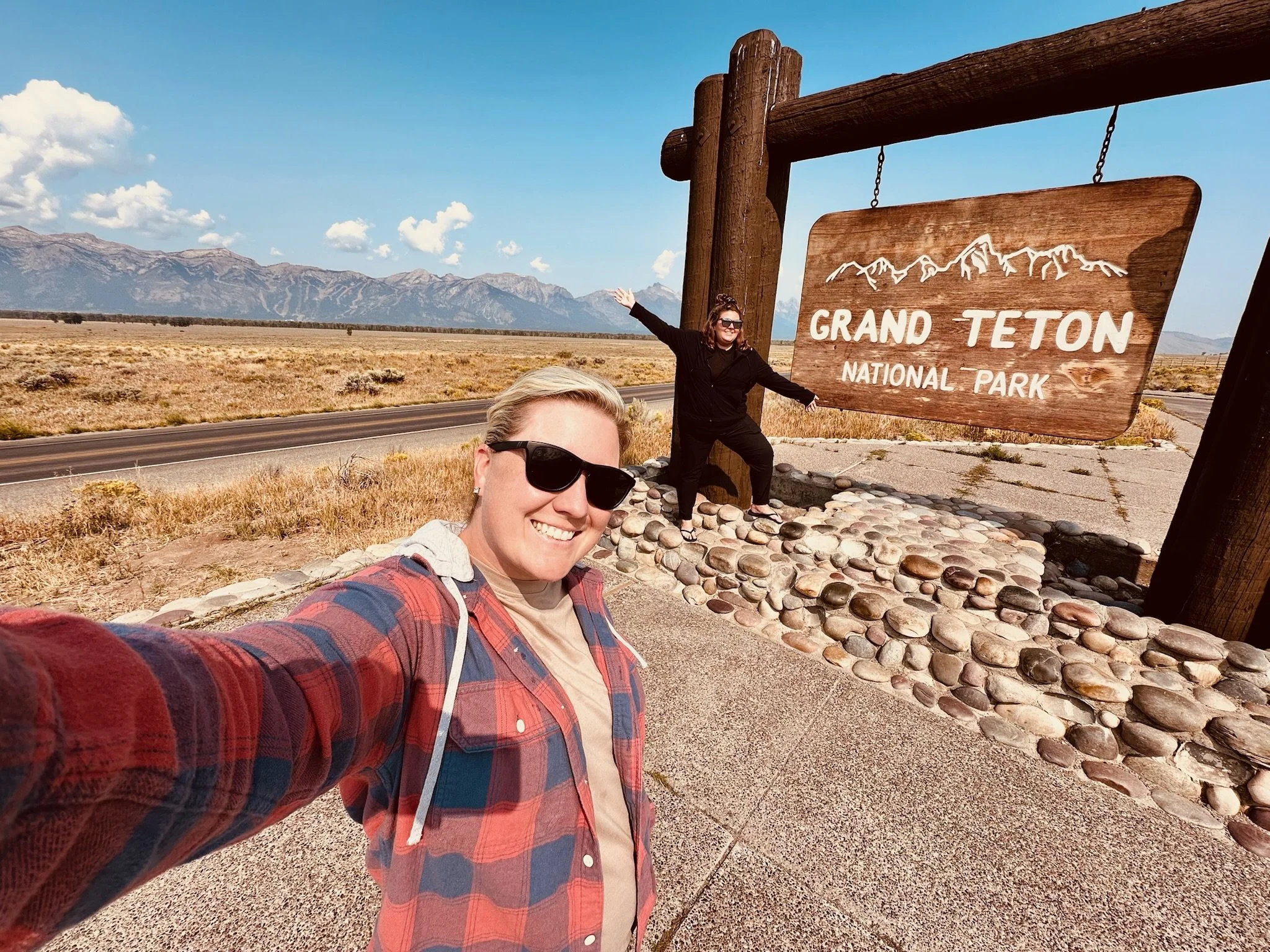 Two smiling women taking a selfie at Grand Teton National Park sign, with mountains and a vast plain in the background. One woman is wearing sunglasses and a plaid shirt, the other is gesturing joyfully.
