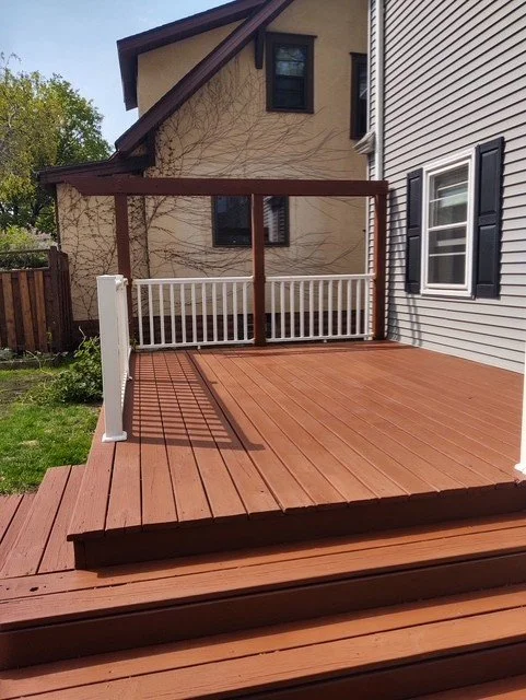 Brown deck with white railing beside gray house.  Tan stucco house in background