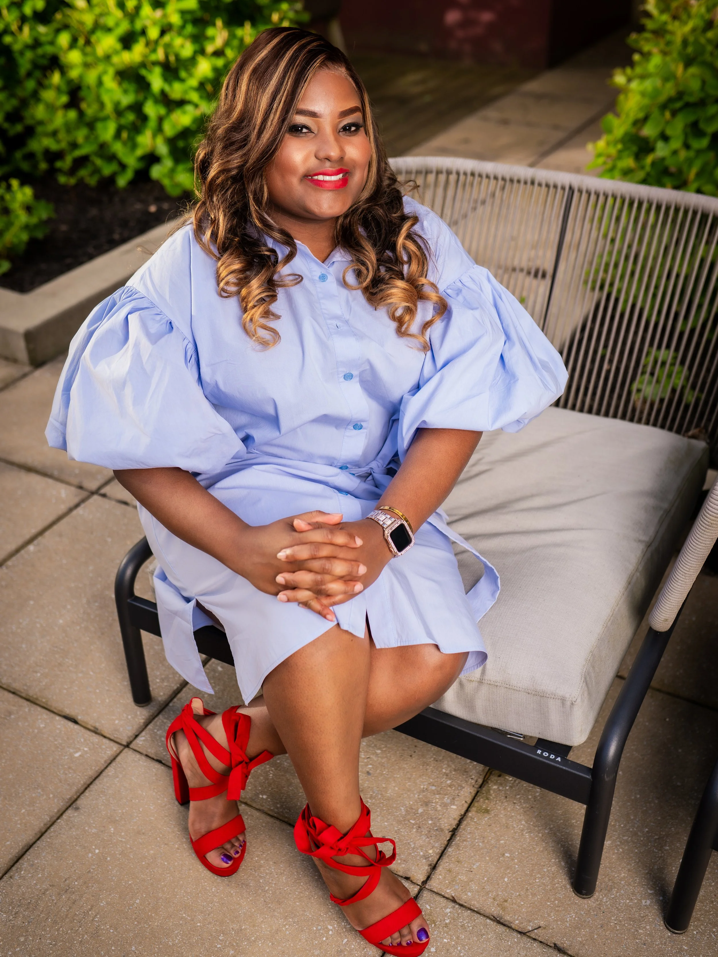 A woman sitting on an outdoor bench, wearing a light blue dress, red high-heeled sandals with ribbon straps, and a smartwatch. She has long, curly hair and is smiling at the camera, with bushes and a paved area in the background.