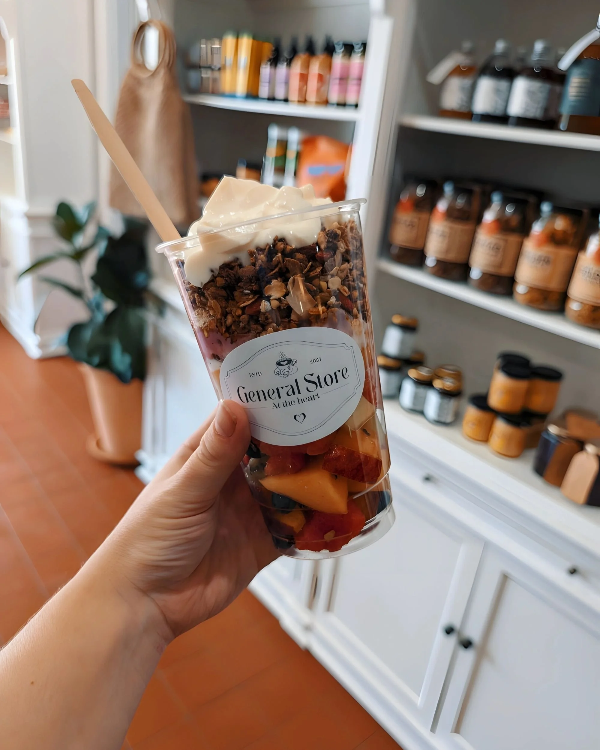 A hand holding a fruit and granola parfait in a clear cup inside a general store. The parfait contains layers of fresh fruit, granola, and yogurt, with a wooden spoon in the cup. Shelves with various products are visible in the background.
