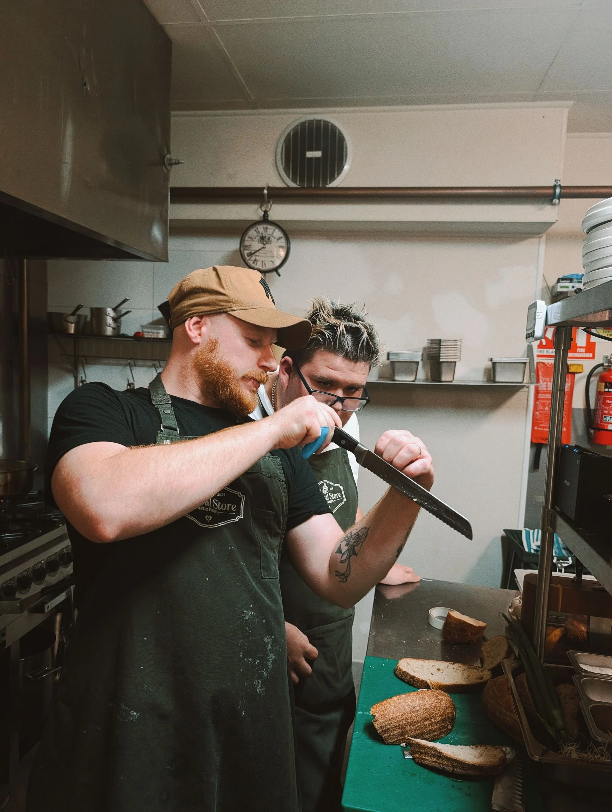 Two men wearing green aprons in a kitchen, one holding a bread knife and the other watching. Sliced bread on a cutting board. Shelves with kitchen supplies in the background.