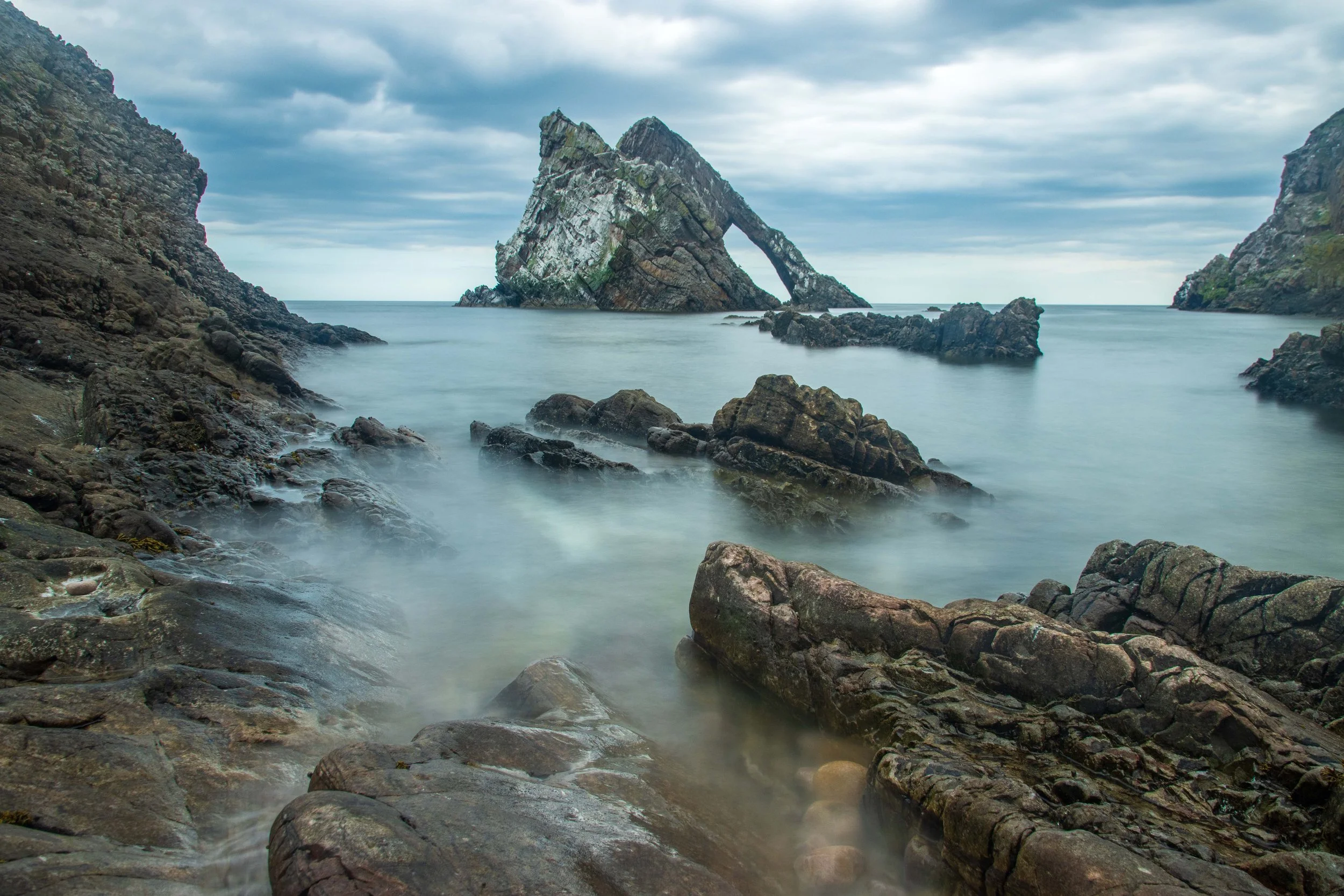 Scenic coastal landscape featuring a large, uniquely shaped rock formation rising from the water, surrounded by smaller rocks and cliffs under a cloudy sky.