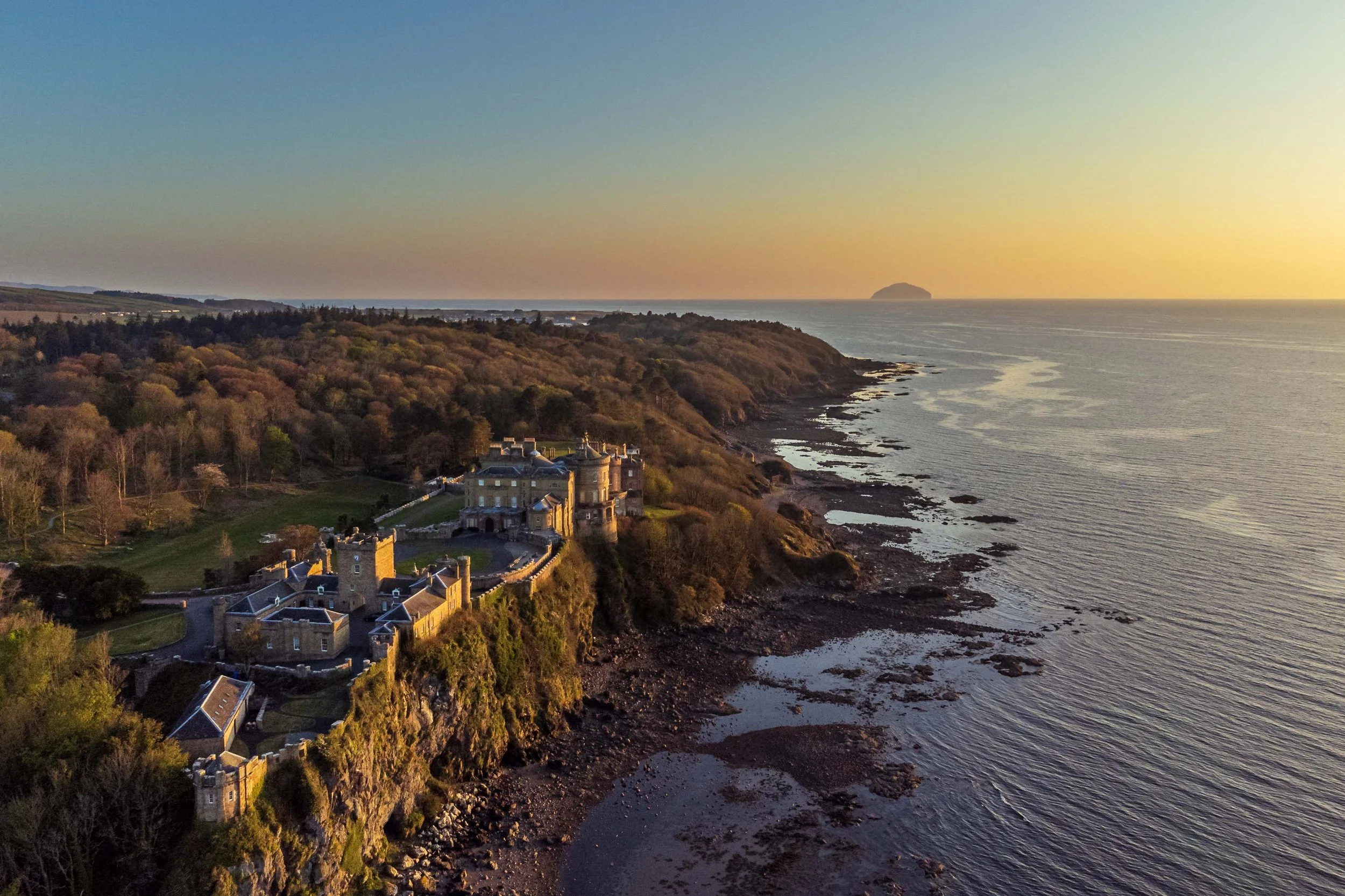 Aerial view of a historic castle on a rocky cliff overlooking the ocean at sunset, with a wooded landscape behind.