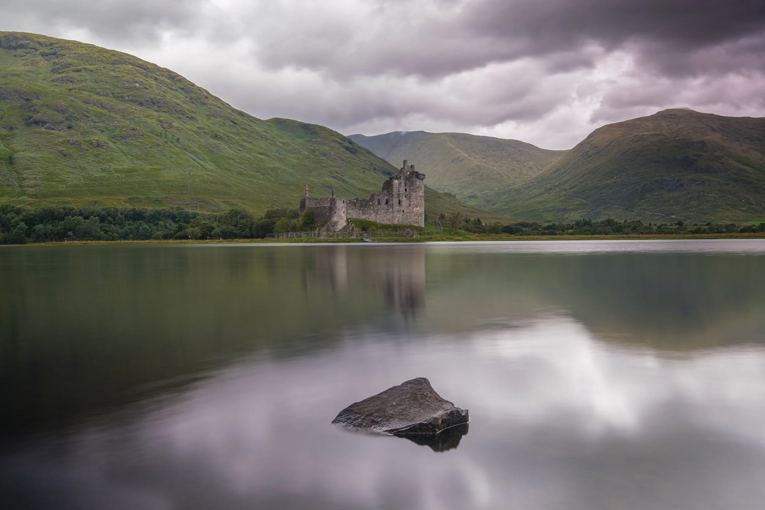 An ancient castle on a lakeshore surrounded by green hills and mountains under a cloudy sky.