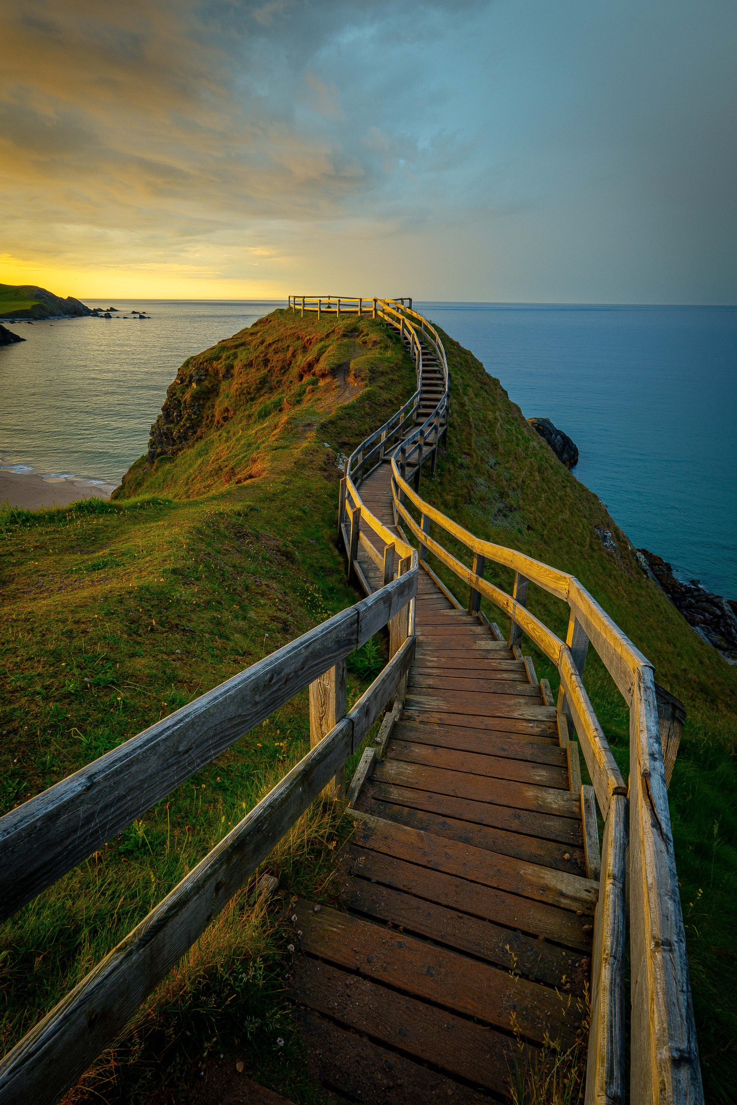 A wooden walkway winding up a grassy hill overlooking the ocean at sunset or sunrise with cloudy sky.