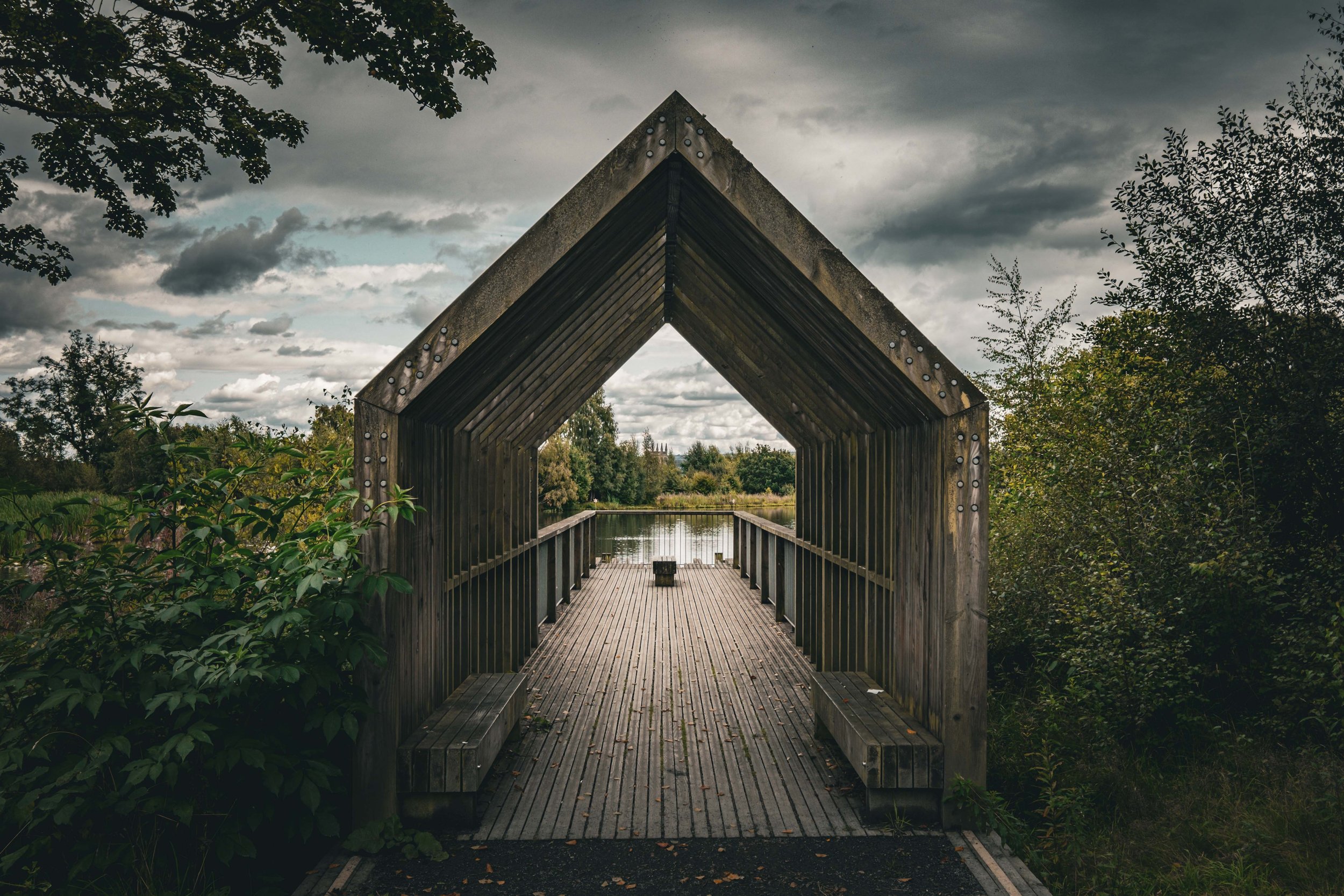 A wooden covered bridge over a waterway with trees and cloudy sky in the background.