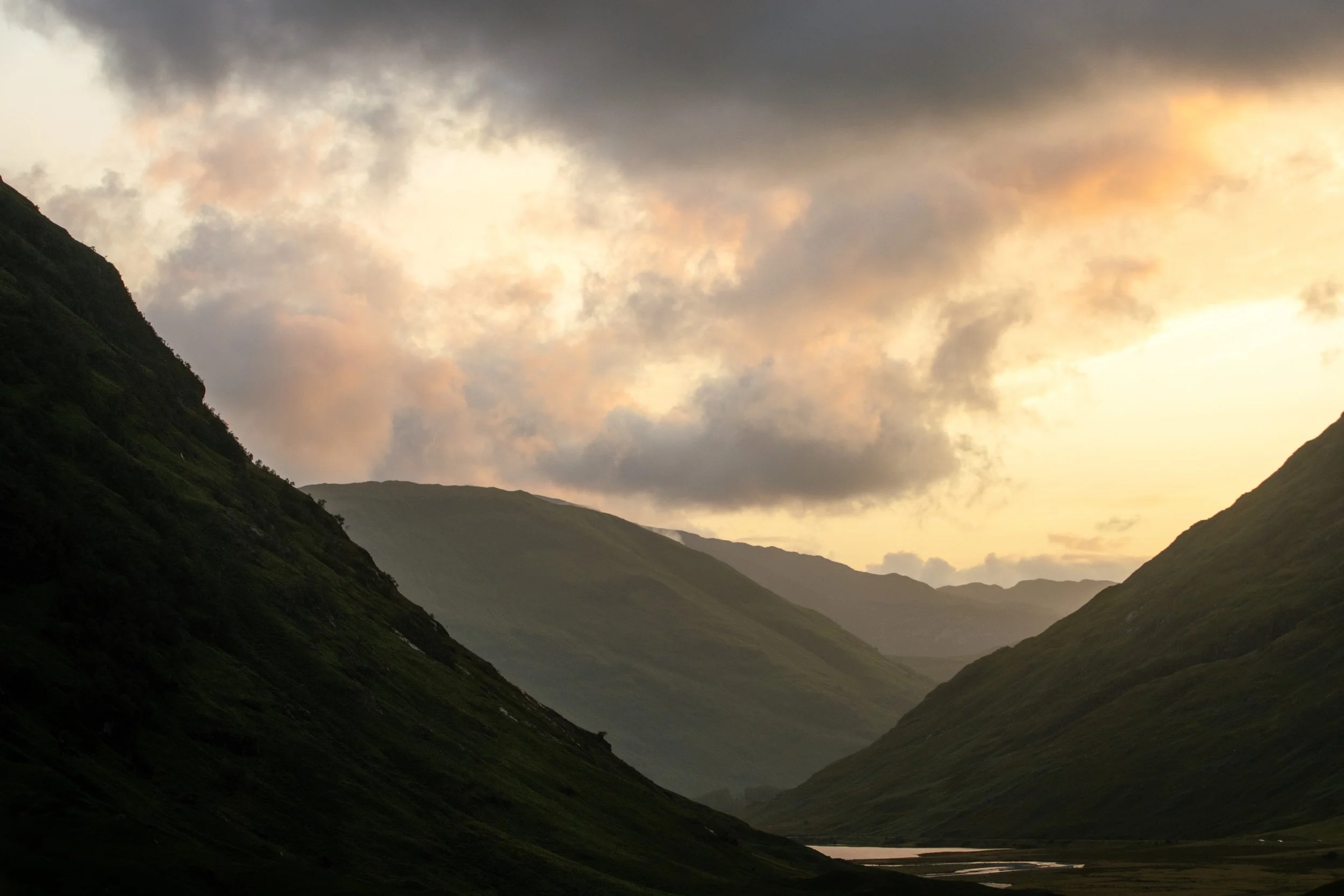 A scenic view of green mountains with a river at the bottom, under a cloudy sky with the sun setting or rising.