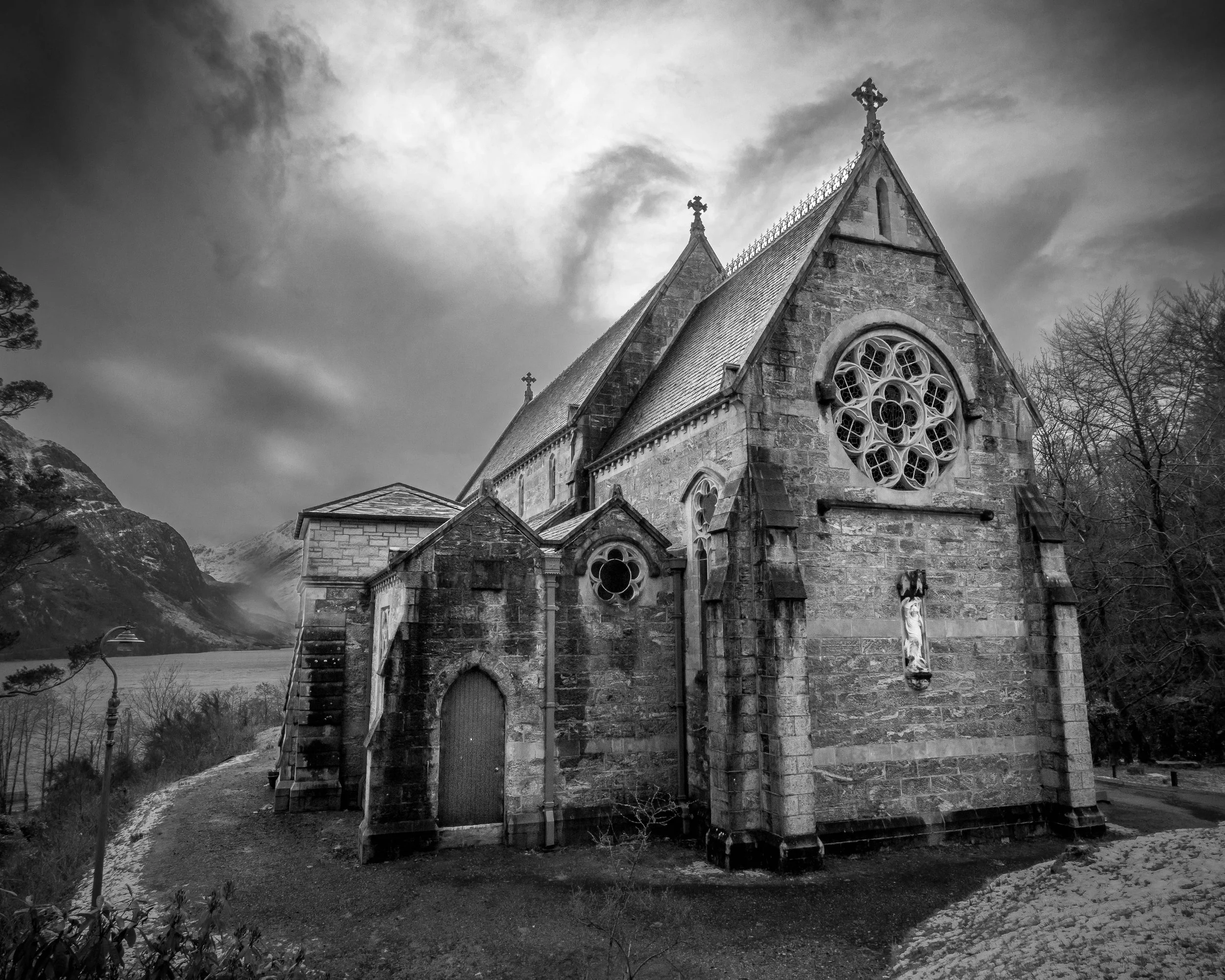 A black and white photo of an old stone church with stained glass windows, set in a mountainous landscape with cloudy skies.