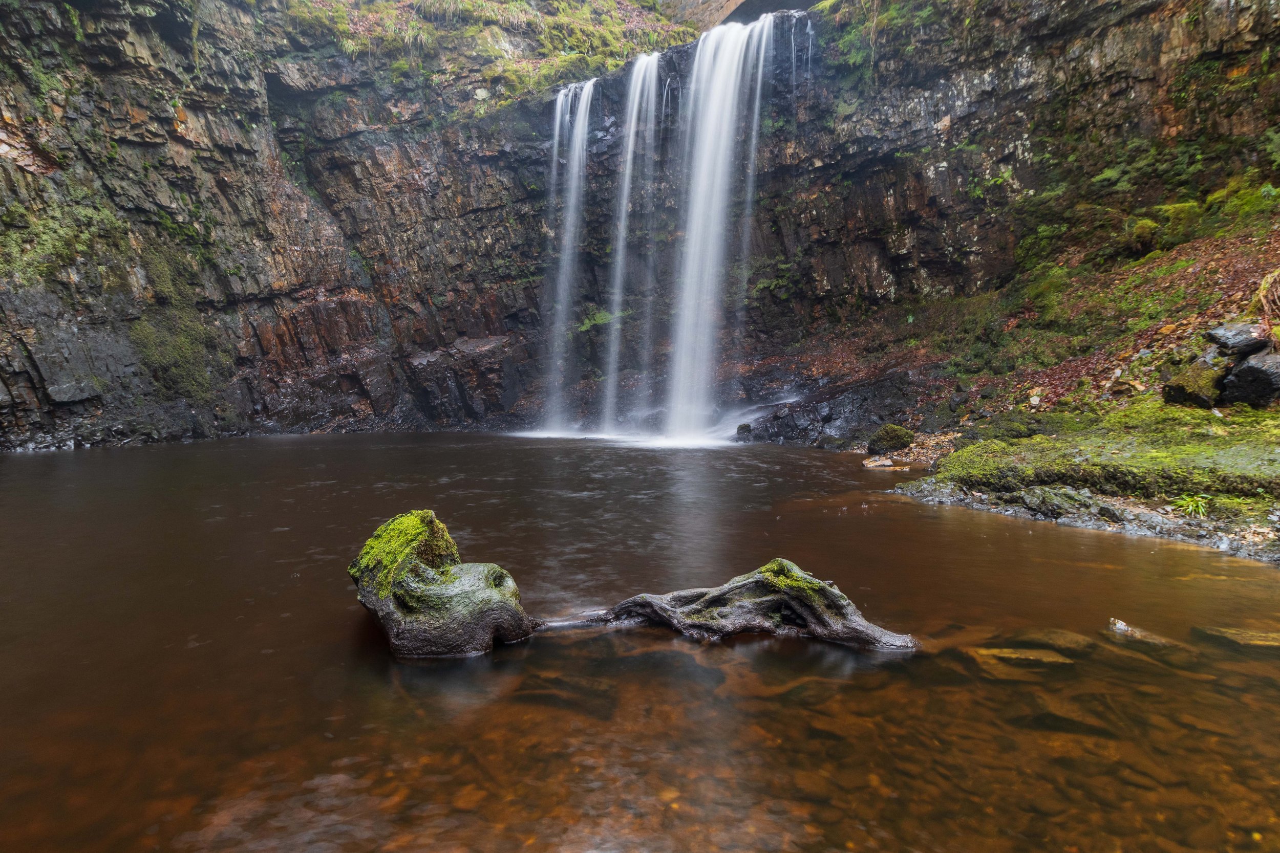 A waterfall flowing into a dark pool surrounded by moss-covered rocks and green foliage.