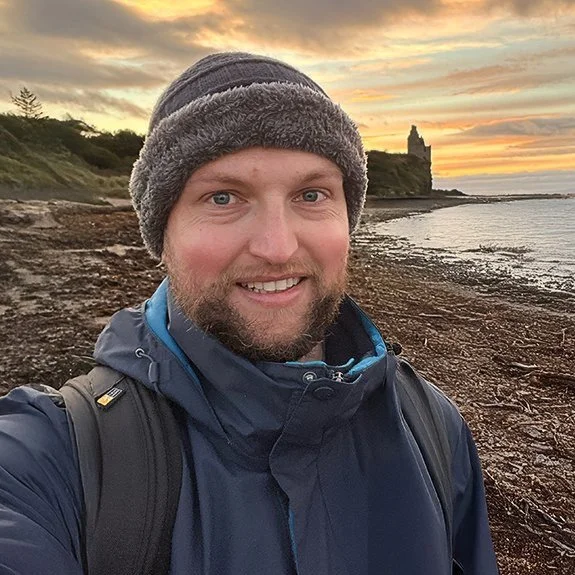 Man taking a selfie at the beach during sunset, wearing a winter hat and jacket, with a castle-like structure on a hill in the background.