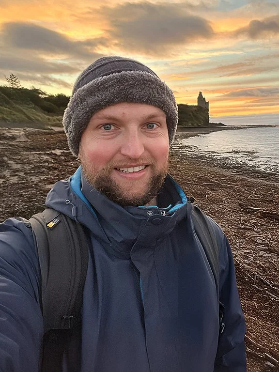 A man taking a selfie on a rocky beach at sunset, wearing a gray beanie, a navy jacket, and a backpack.
