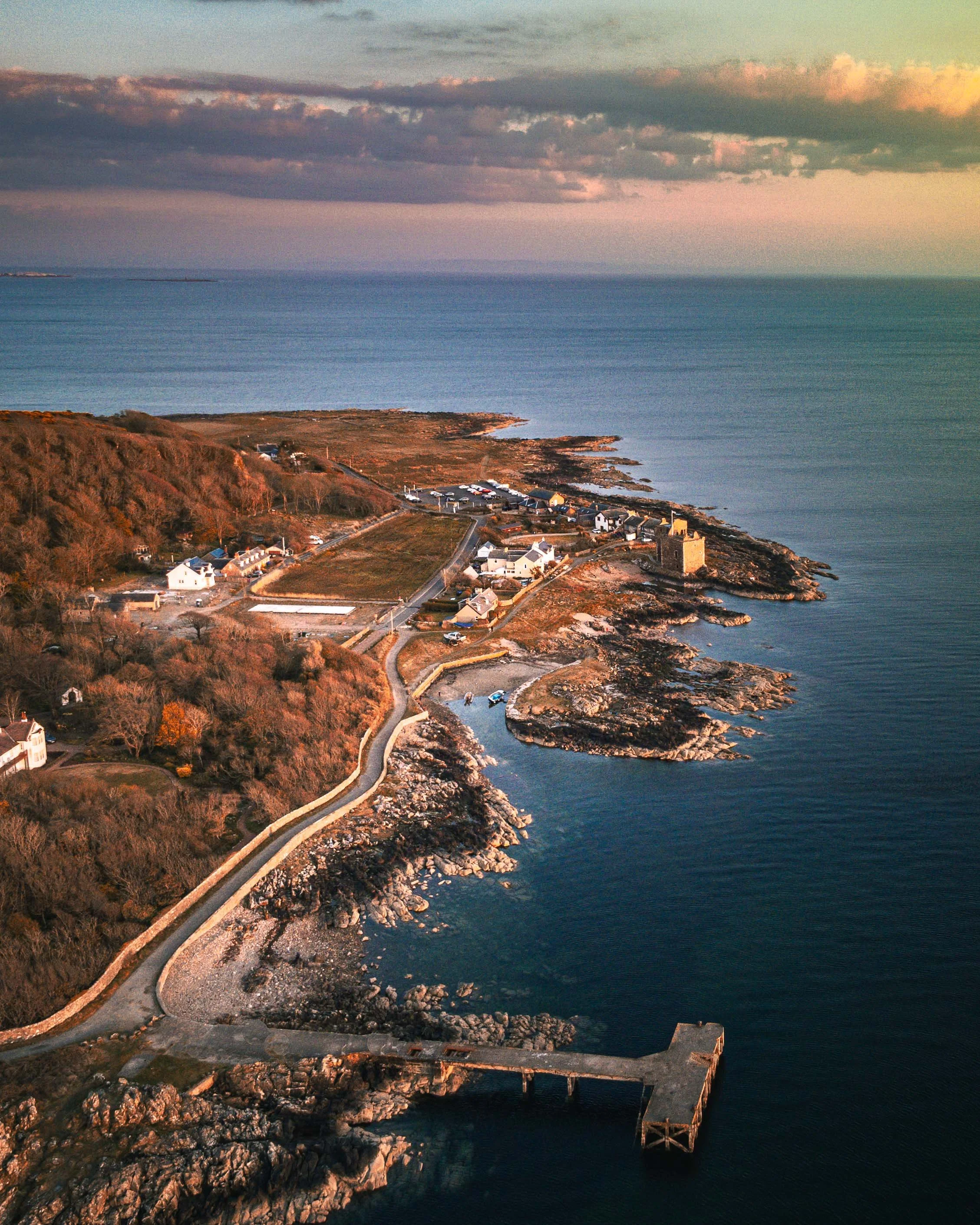 Aerial view of a coastal village with houses, a lighthouse, a pier, and rocky shoreline at sunset, overlooking the ocean with cloudy sky.