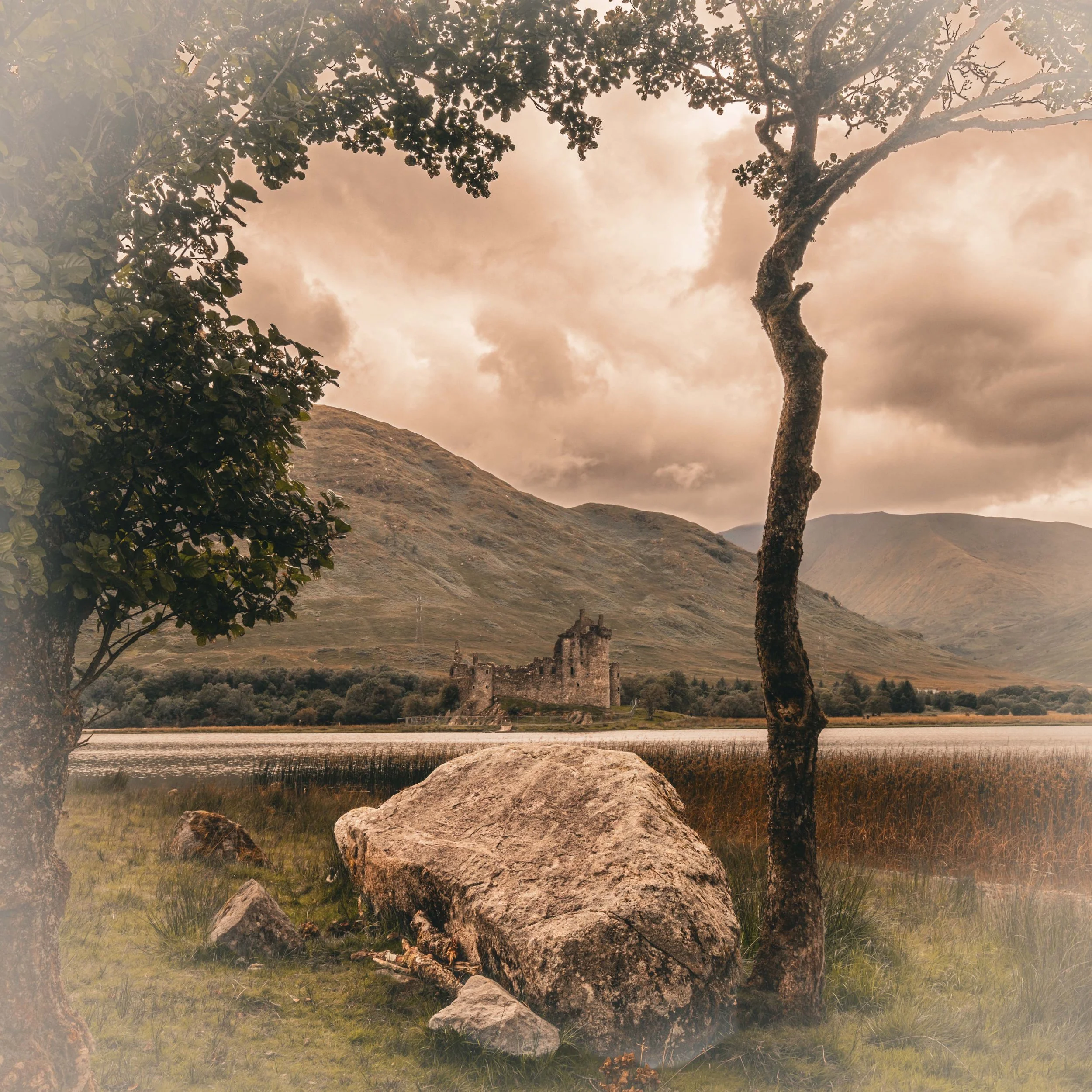 A scenic view of a lake with a large rock in the foreground, two trees on either side, a castle in the middle distance, and mountains in the background under a cloudy sky.