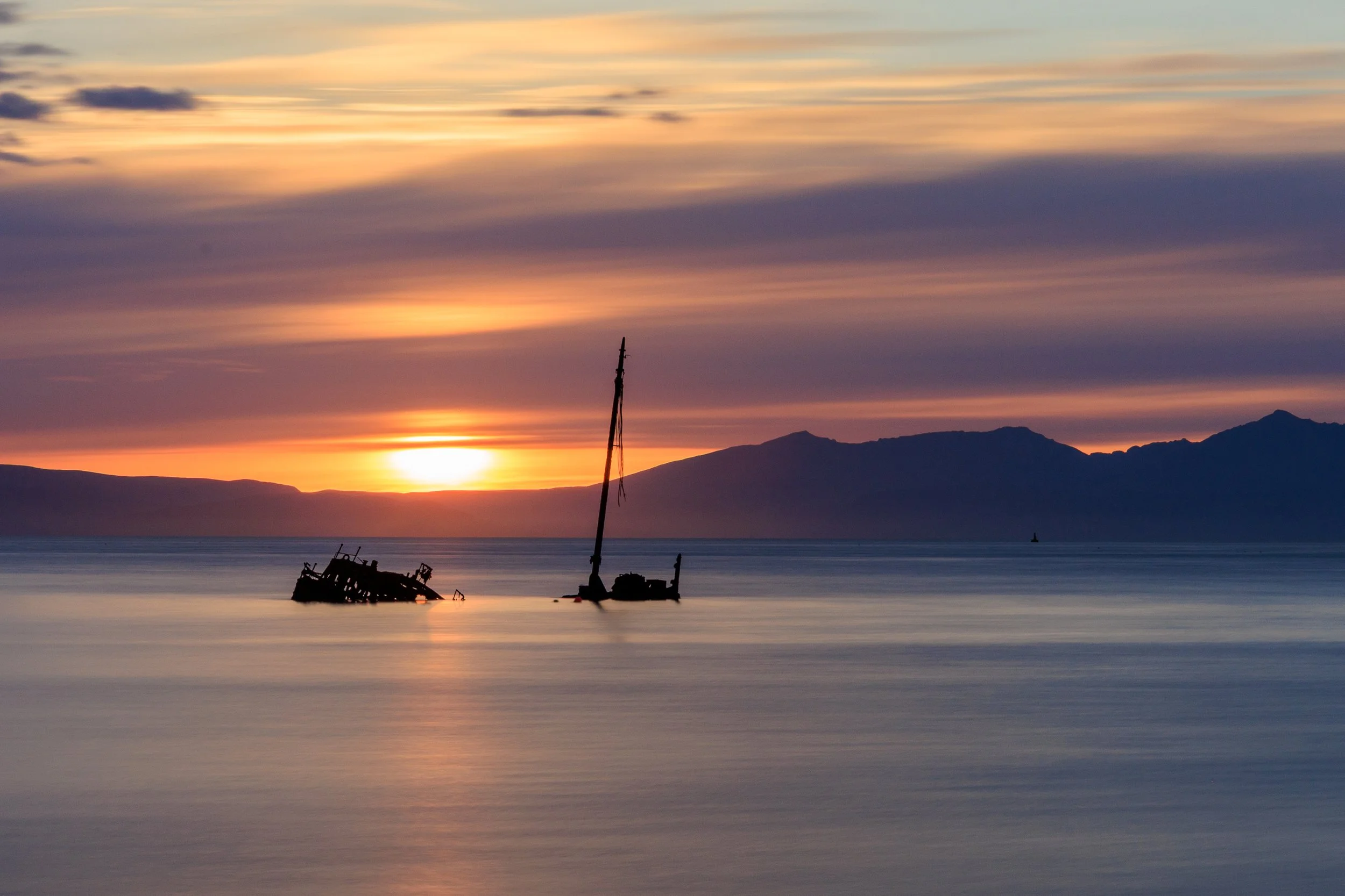 Sunset over calm water with two wrecked ships and mountains in the background.