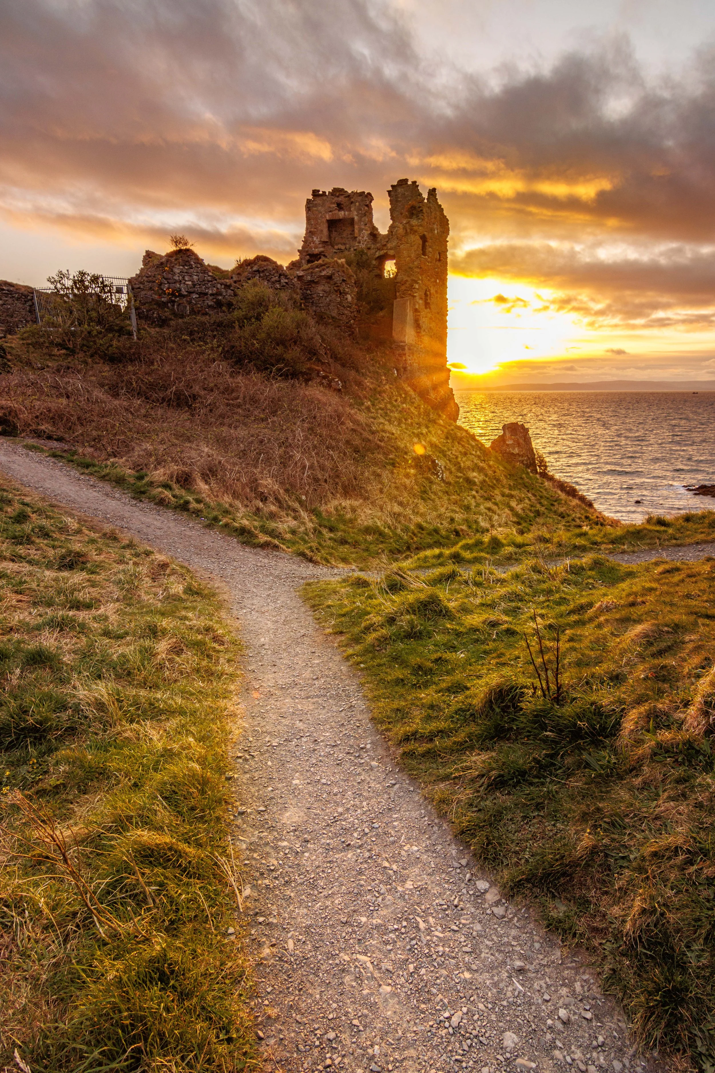 A dirt pathway leading towards the ruins of a castle on a hill, with the sunset and ocean in the background.