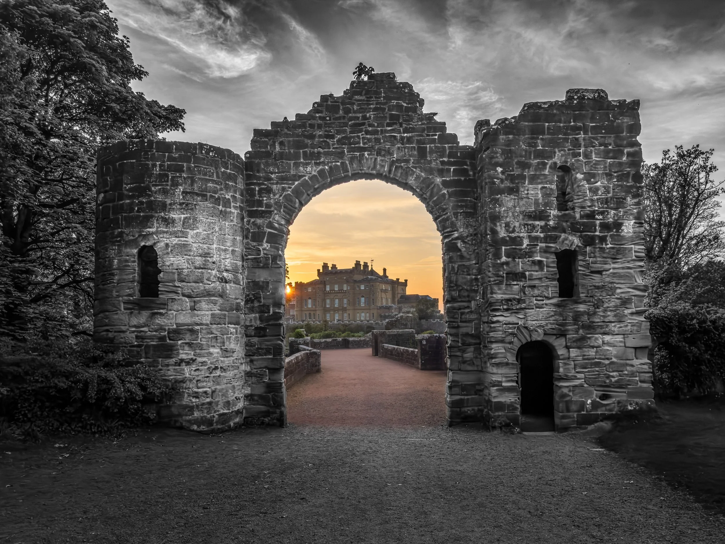 Stone archway ruins with a castle in the background at sunset, black and white with a hint of color in the sky and distant building.