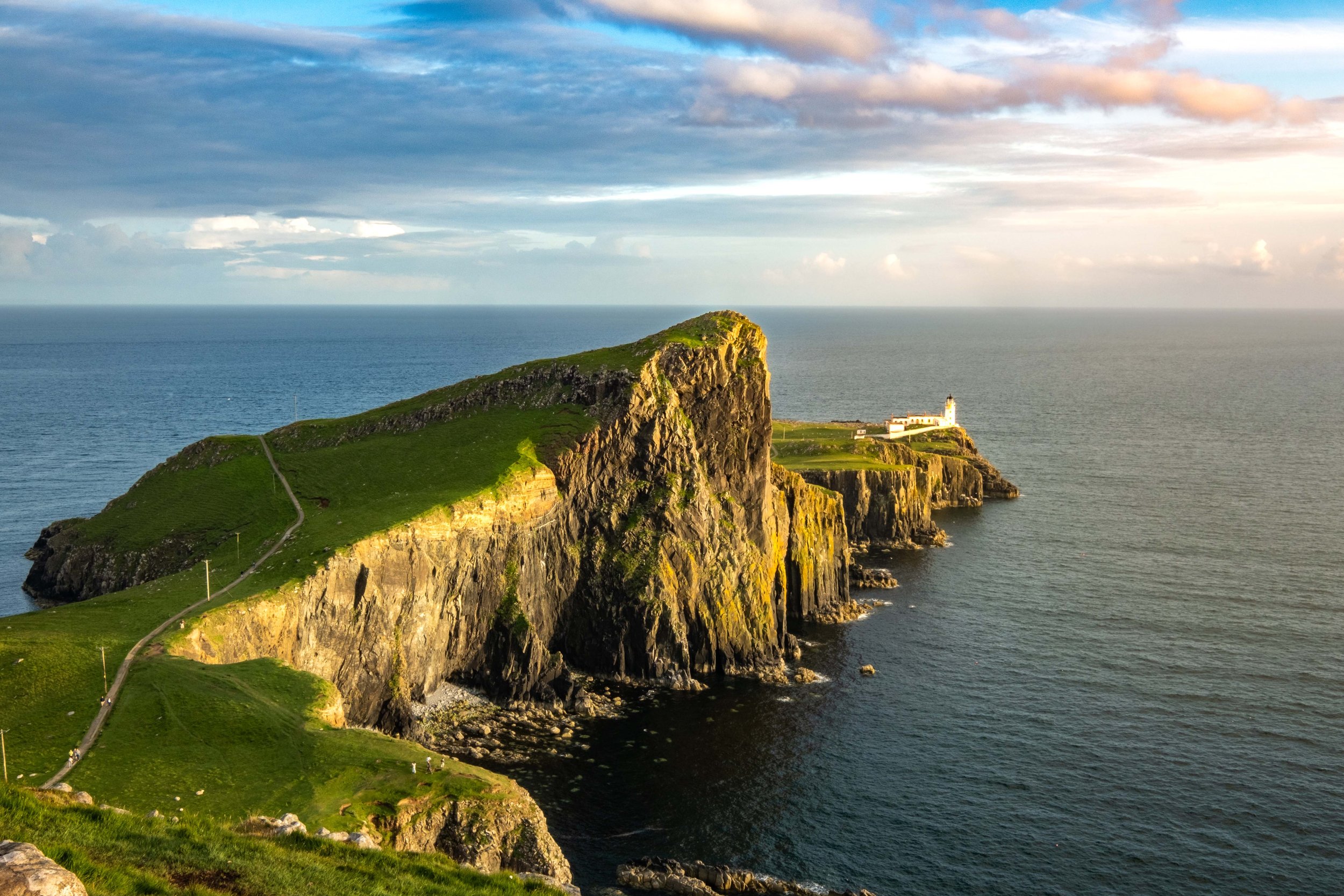 A coastal landscape with green cliffs overlooking the ocean, a winding path, and a small white lighthouse in the distance at sunset.