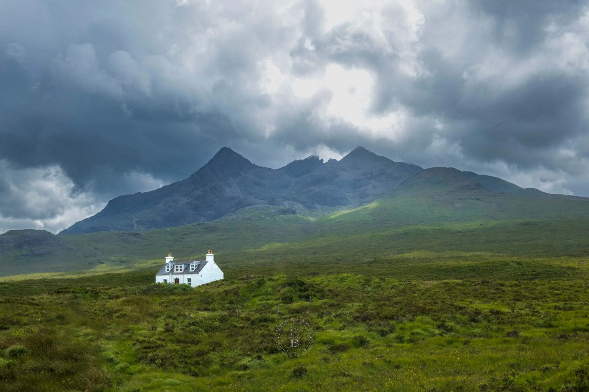 A white house with dark roof and chimneys surrounded by green grass and shrubs, with mountains and cloudy sky in the background.