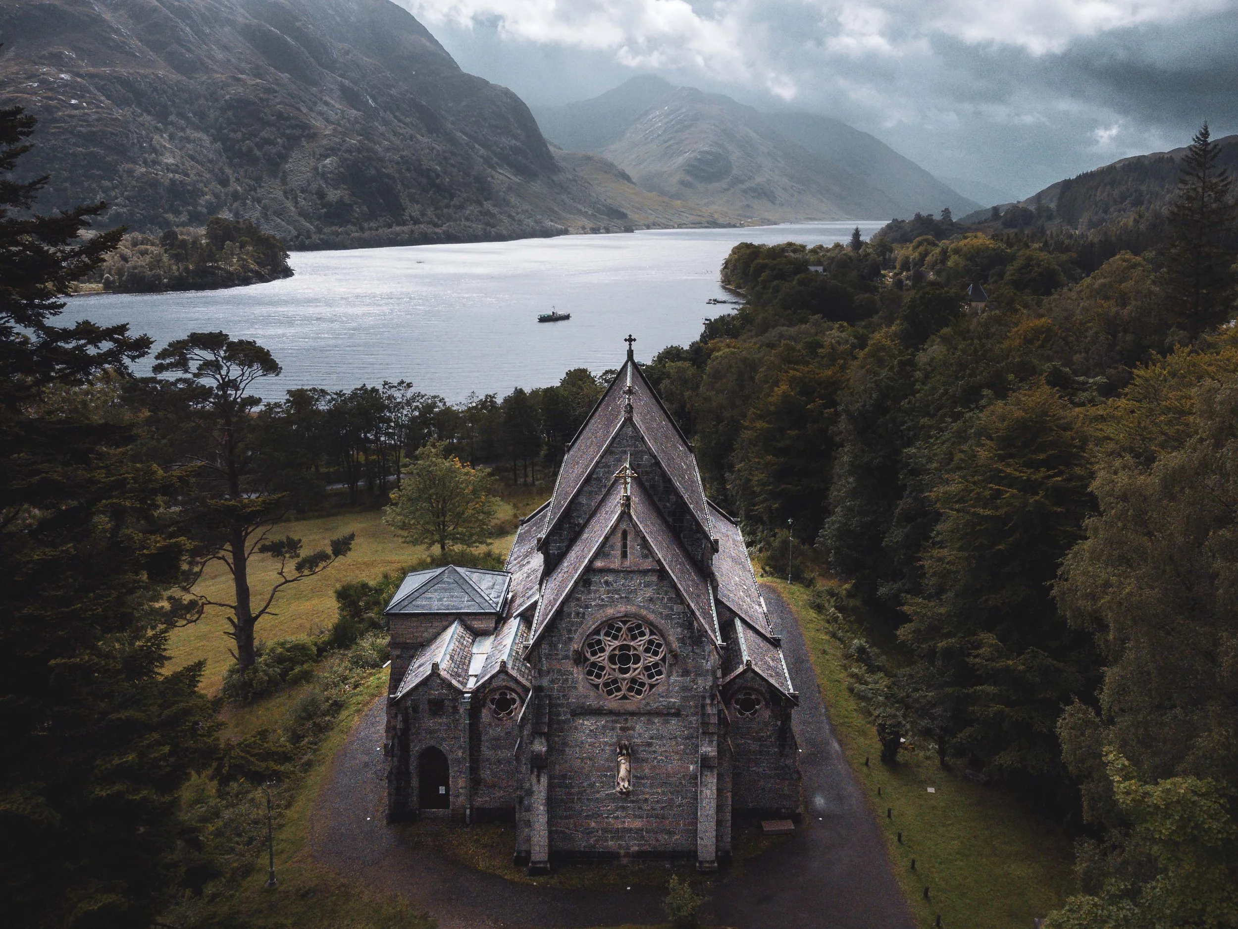 A stone church surrounded by trees, with a lake and mountains in the background on a cloudy day.