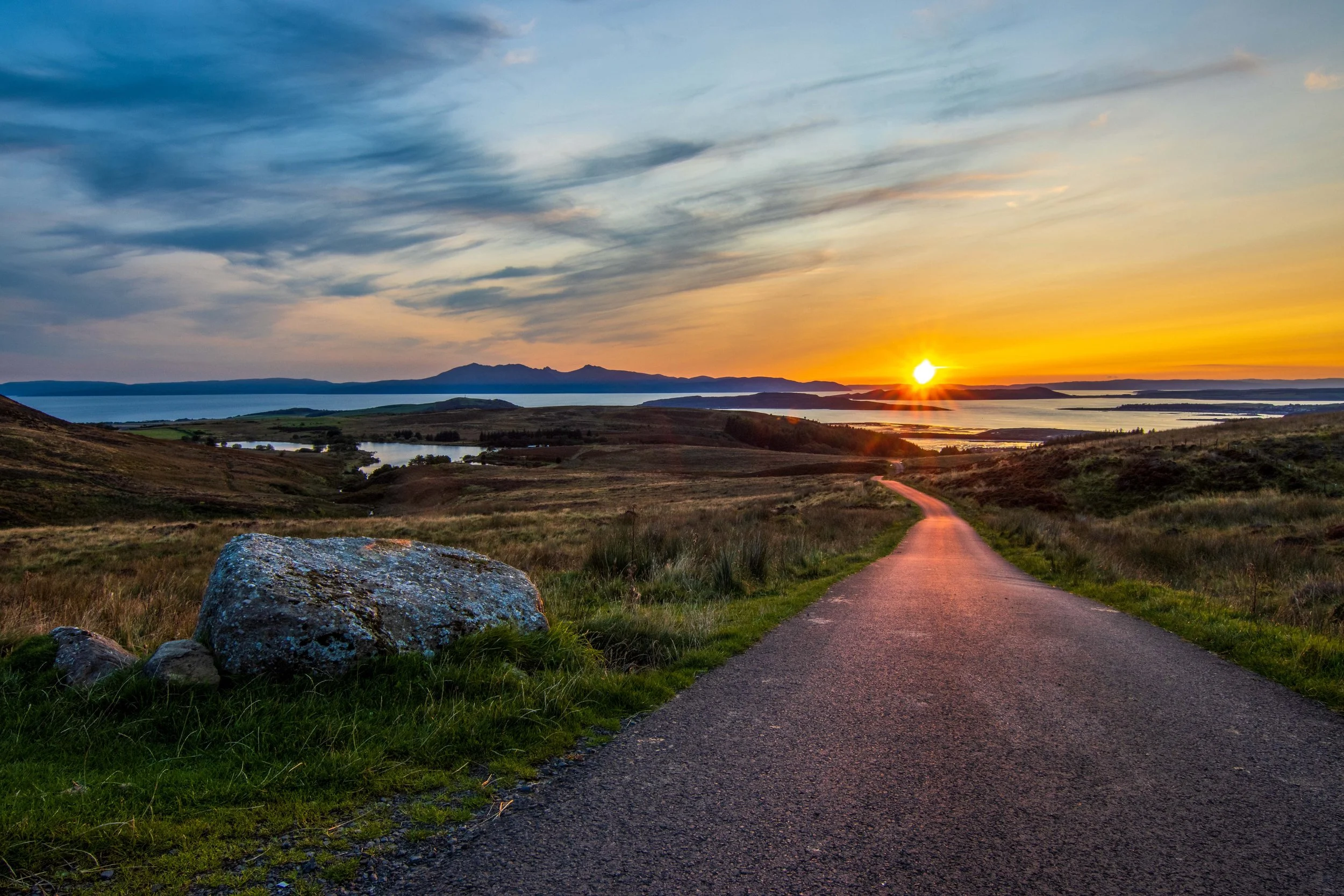 A scenic landscape at sunset with a winding road leading to a lake, rolling hills, and mountains in the distance, under a sky with colorful clouds.