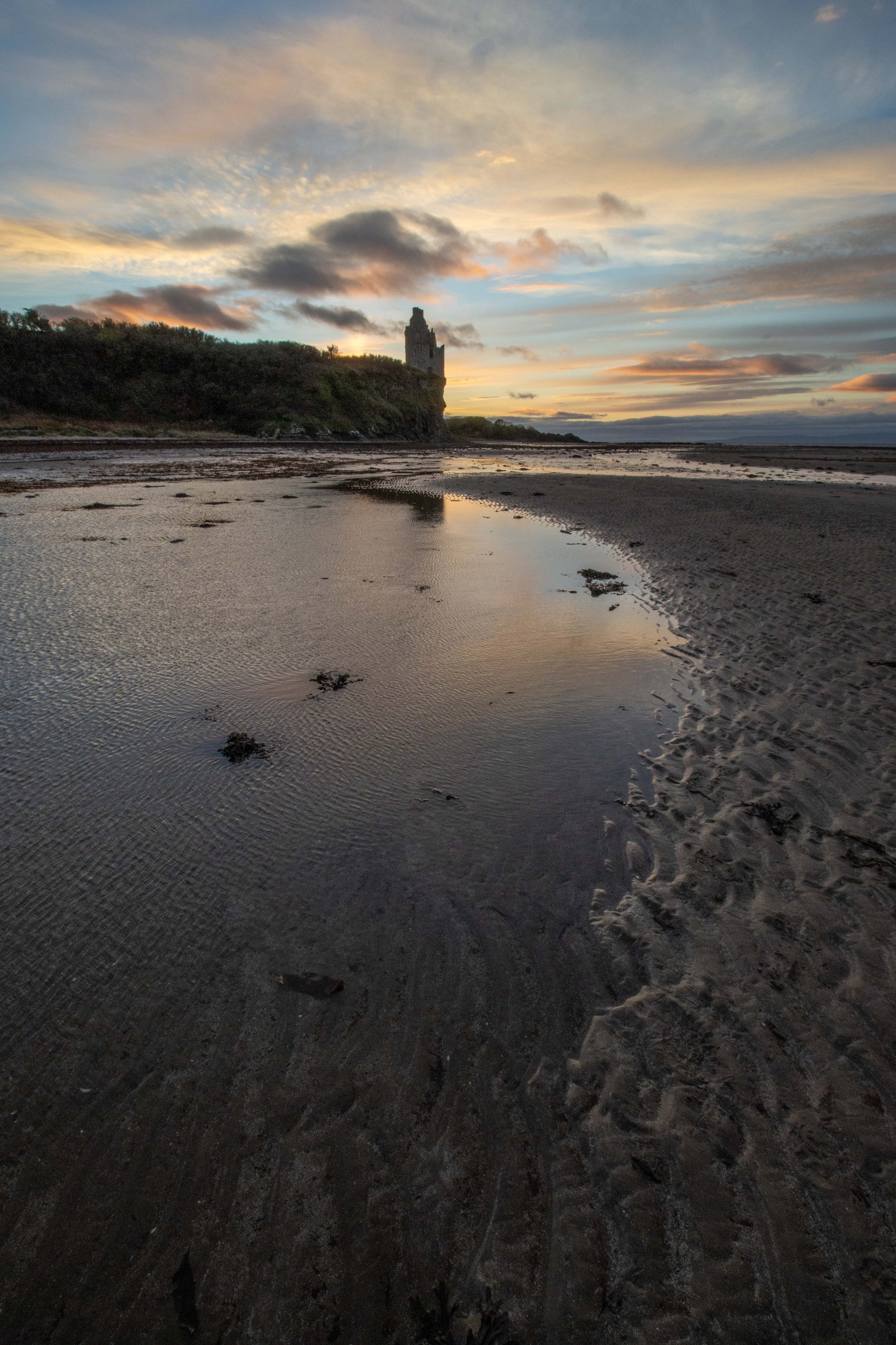 Sunset over sandy beach with a small stream of water, and ruins of a castle or tower in the distance.