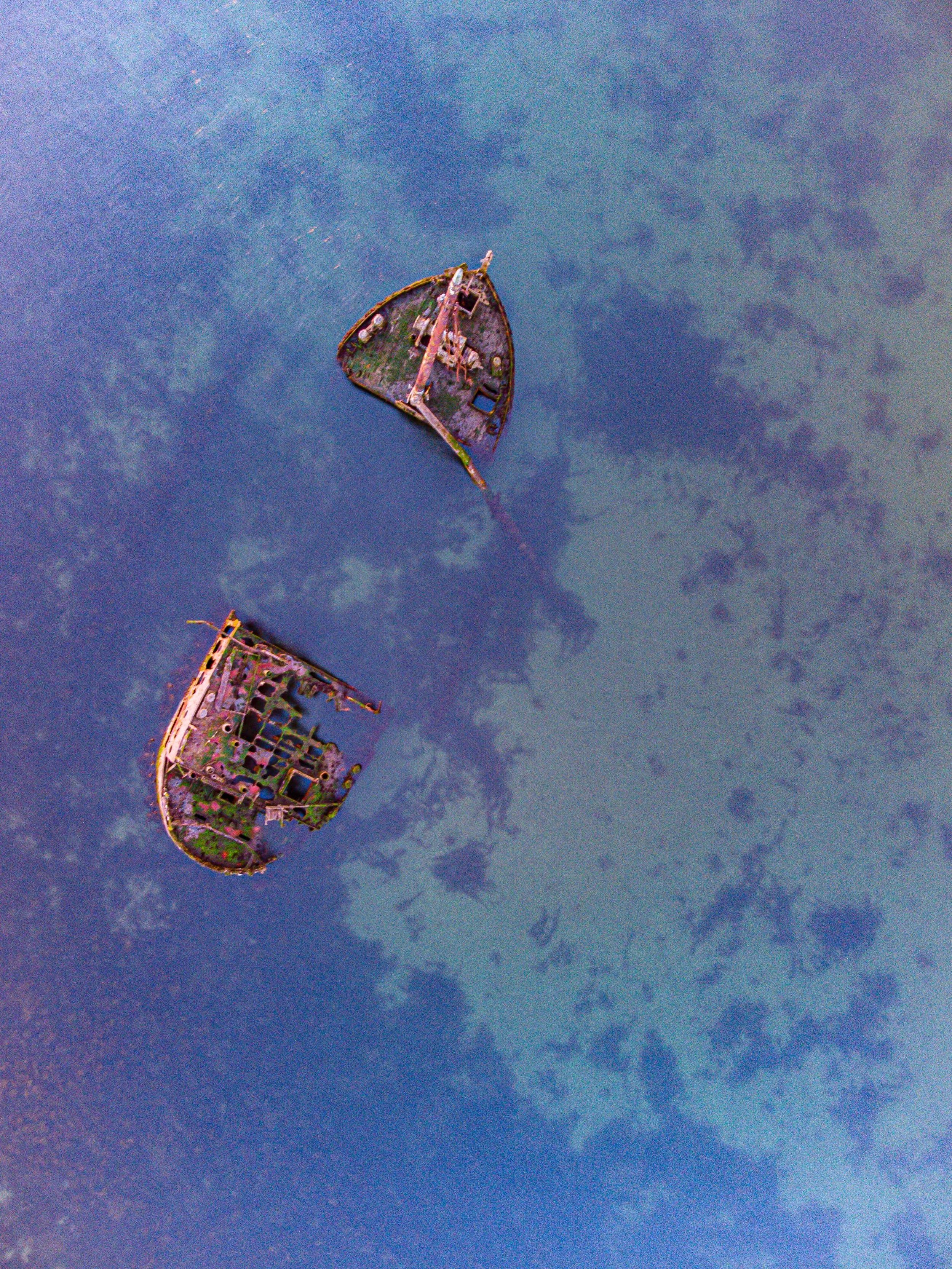 Birds-eye view of two submerged, dilapidated shipwrecks in calm water.