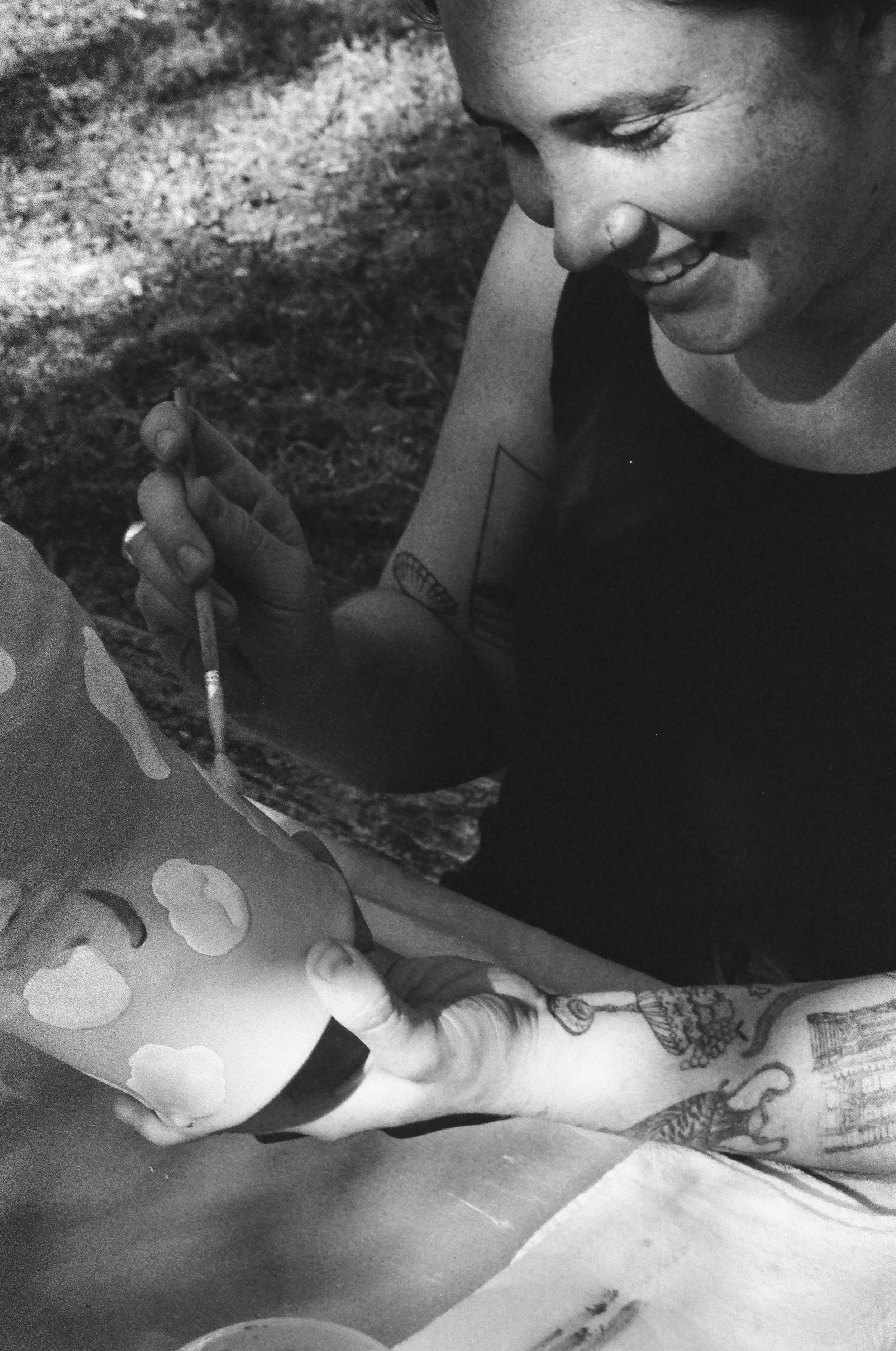 Person smiling while painting a large object with a small brush, with a tattooed arm visible.