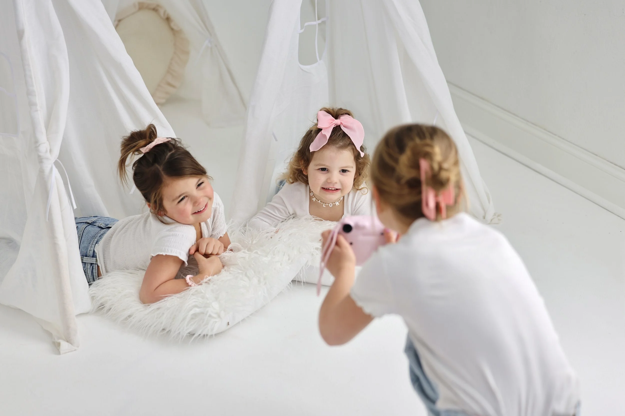 Three young girls with curly hair, wearing white shirts, are inside a white tent-like structure with flowing fabric. One girl is lying on her stomach on a fluffy white rug, smiling at the camera. Another girl is also lying on her stomach, propped up 