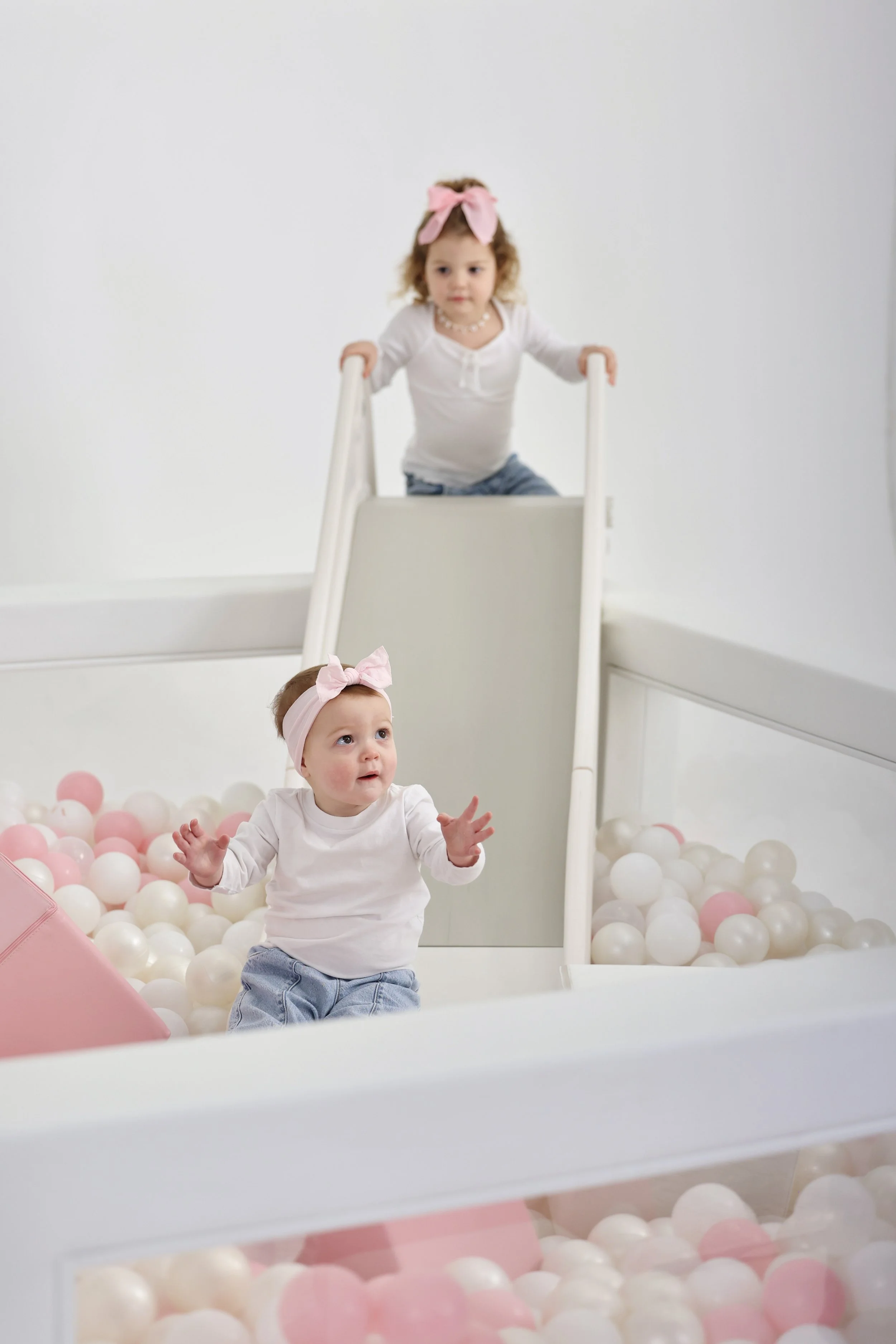 Two young girls with pink bows in their hair playing in a ball pit with a slide, in a white room.