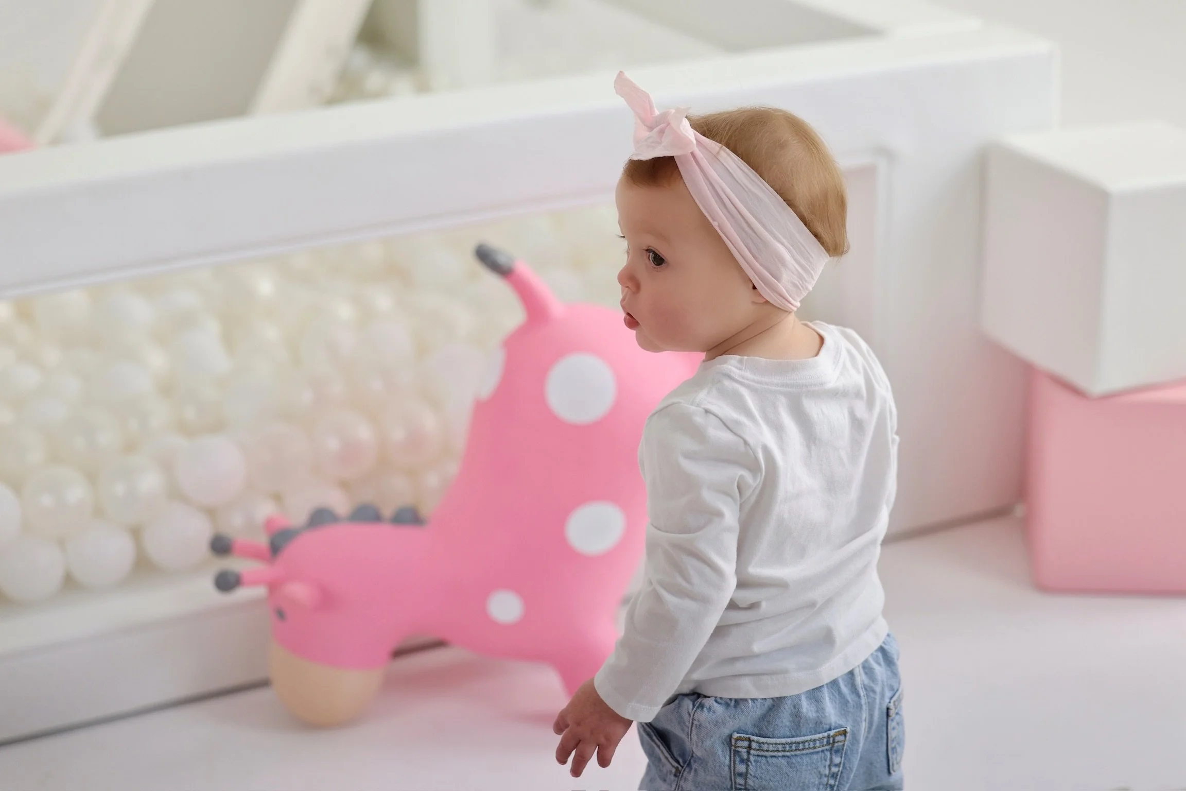 A young girl wearing a pink headband and a white long sleeve shirt stands near a pink inflatable unicorn with white polka dots on it, in a room decorated with pastel colors.