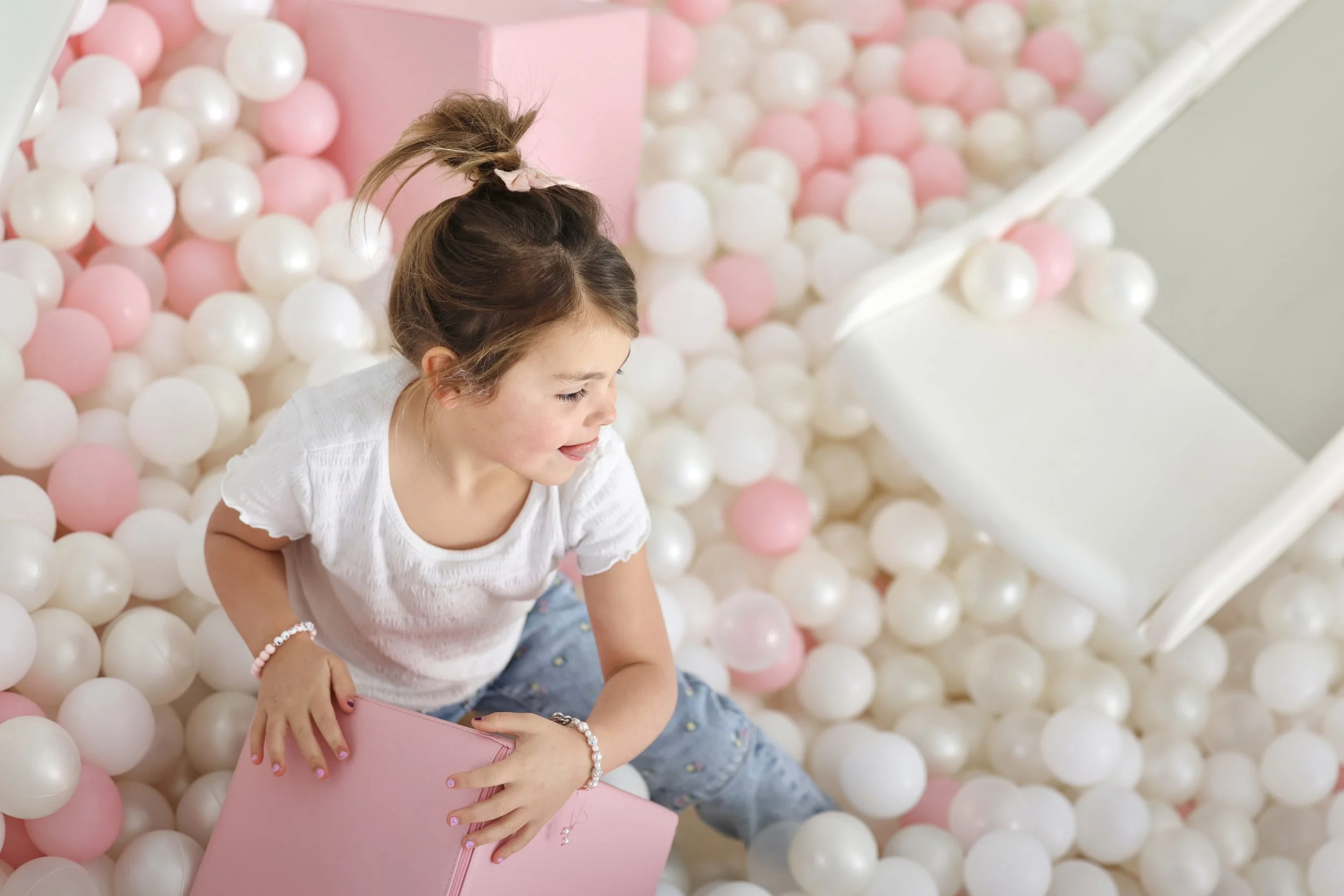 A smiling girl with a white shirt and blue jeans sitting in a ball pit filled with white and pink plastic balls, holding a pink box.