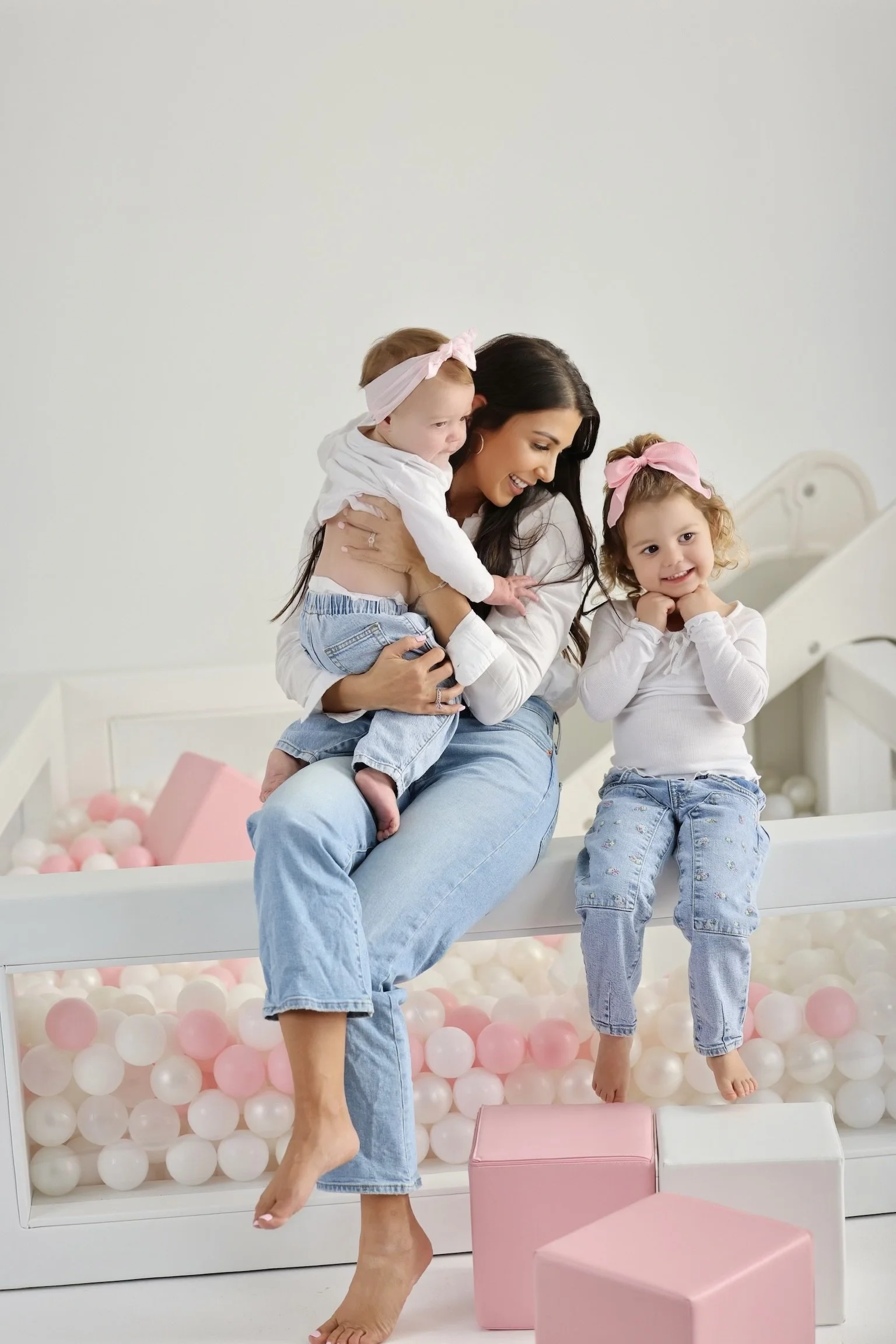A mother and two young daughters sitting on a bed with pink and white balls in a ball pit. The mother is holding one of the daughters, smiling, while the other daughter sits beside them, smiling with hands on cheeks.