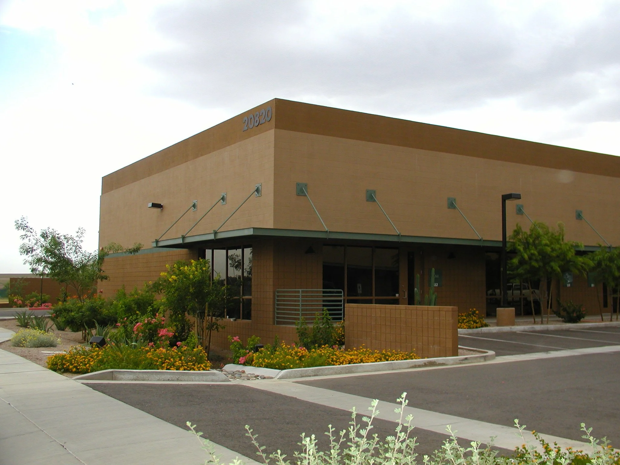 Single-story commercial office building with beige CMU walls, overhangs, and landscaped surroundings including flowers and small trees.