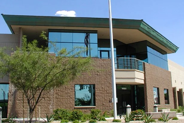 Modern two-story office building with large windows, surrounded by landscaped greenery and a flagpole in front.