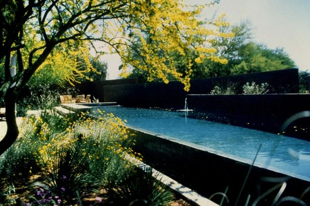 A serene garden scene featuring a long rectangular pond with a small fountain, bordered by lush green plants and yellow flowering trees. In the background, there are outdoor seating arrangements. The sky is clear and blue.