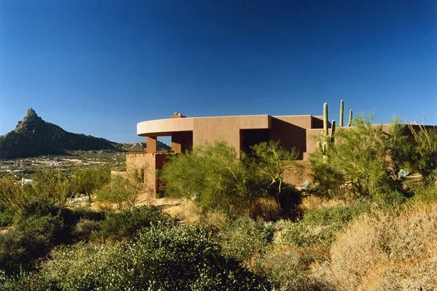 Modern desert home with stone masonry and cacti in the foreground, set against a backdrop of desert hills and blue sky.