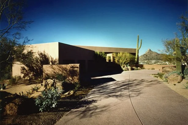 Modern desert home with flat roof surrounded by cacti and desert landscaping. Large driveway leading to the entrance. Clear blue sky in the background.