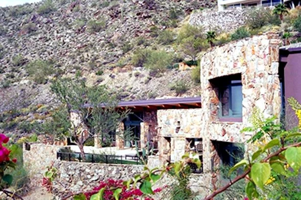 Stone house with large windows integrated into a hillside landscape, surrounded by desert vegetation and flowers.