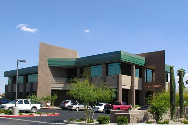 Modern office building with angular design, two stories, large windows, located in a parking lot with several parked cars and desert landscaping including cacti and small trees.