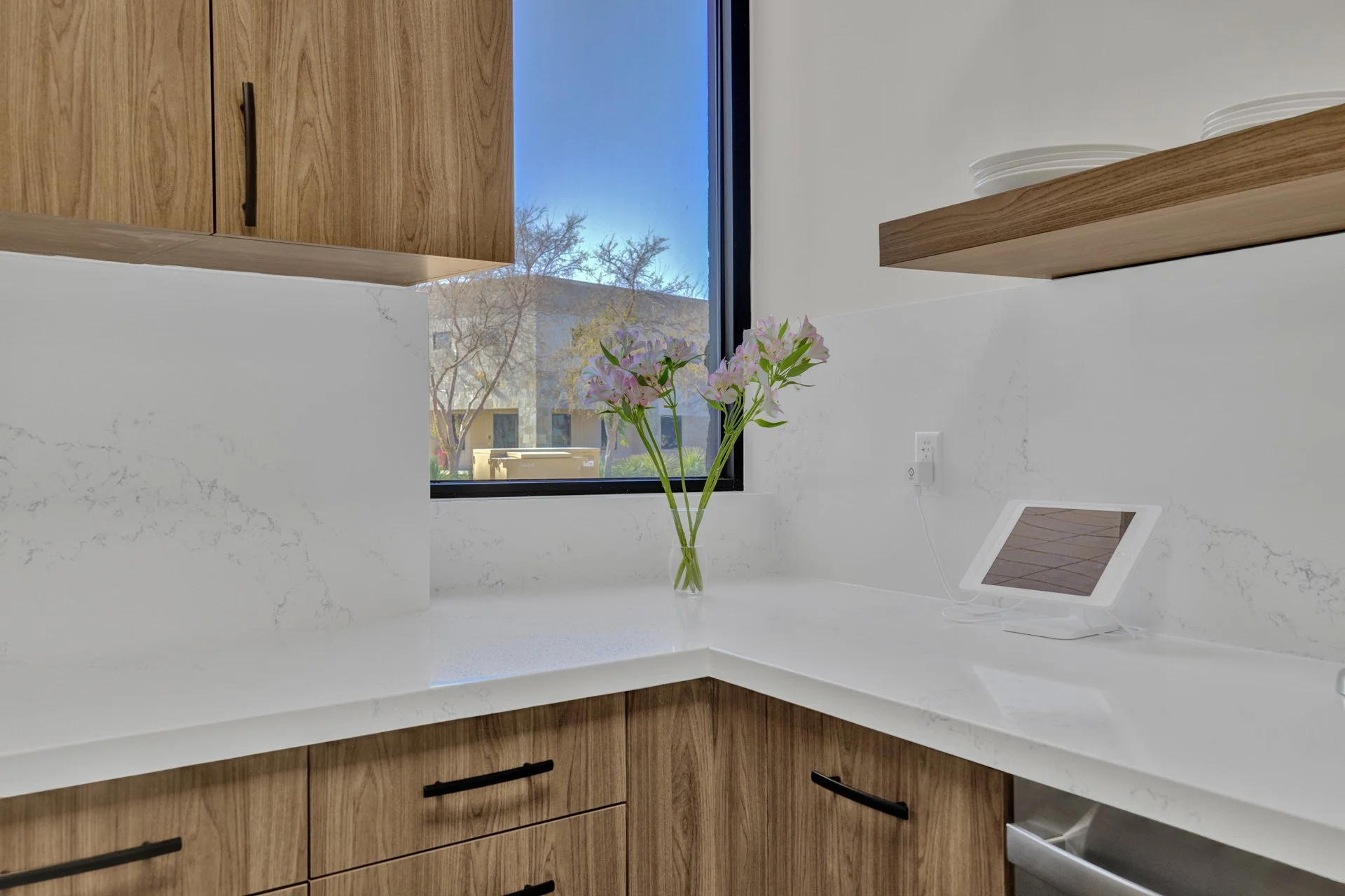 Modern kitchen corner with wooden cabinets, white quartz countertop, vase of pink flowers, and a tablet on a stand near a window.