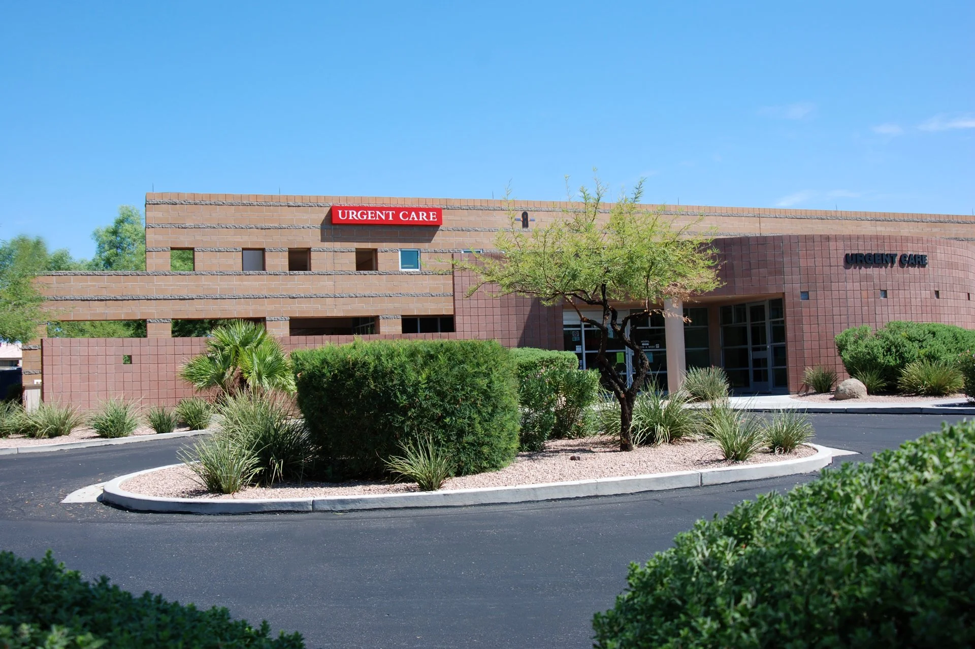 Exterior view of an urgent care building with landscaped shrubs and trees under a clear blue sky.