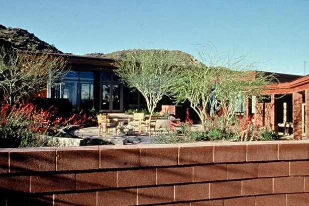 Single-story modern house with large windows, surrounded by desert landscaping and a brick wall in the foreground.