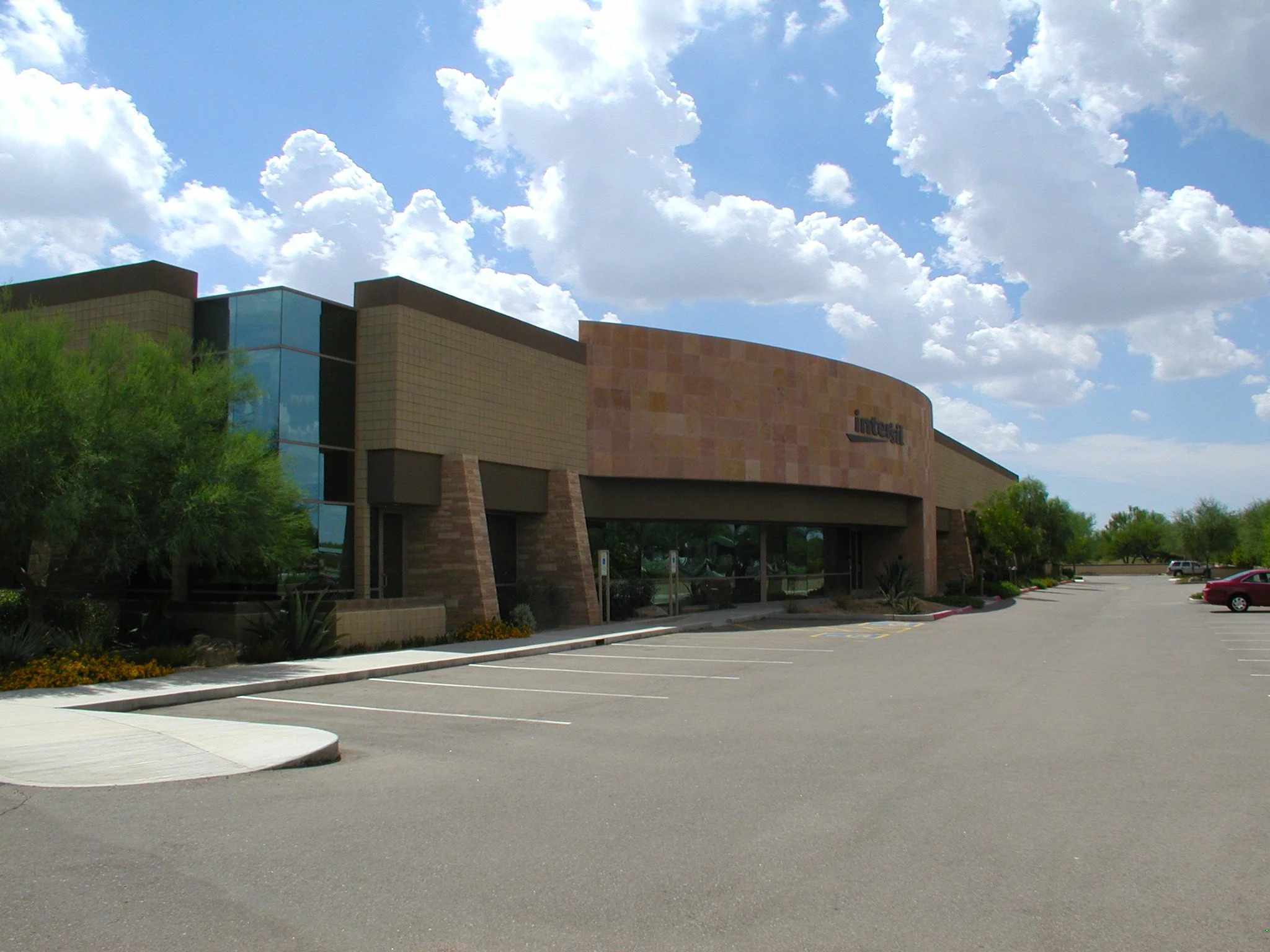 Modern office industrial building with angular design, glass windows, and landscaped surroundings under a clear blue sky with puffy clouds.