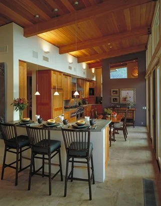 Modern kitchen with a wooden ceiling, bar stools, dining table, and pendant lighting.