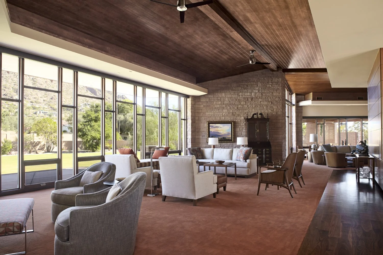 Spacious modern living room with large windows, neutral-toned furniture, and a high wooden ceiling. The room has multiple seating areas with armchairs, sofas, and coffee tables, providing a view of a green outdoor area through floor-to-ceiling window