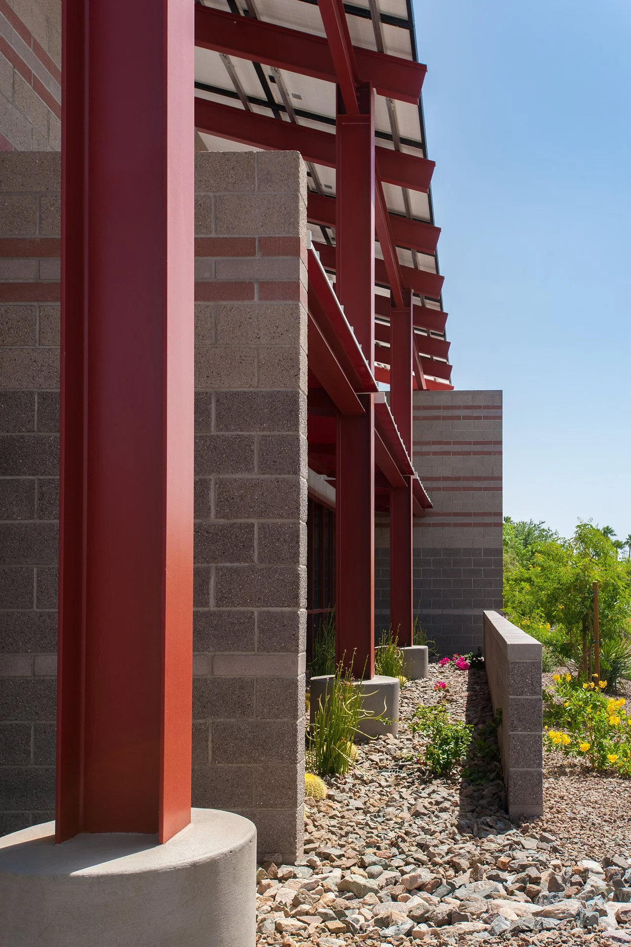 Modern architectural structure with red steel beams, gray brick walls, and landscaped garden with rocks and flowers.
