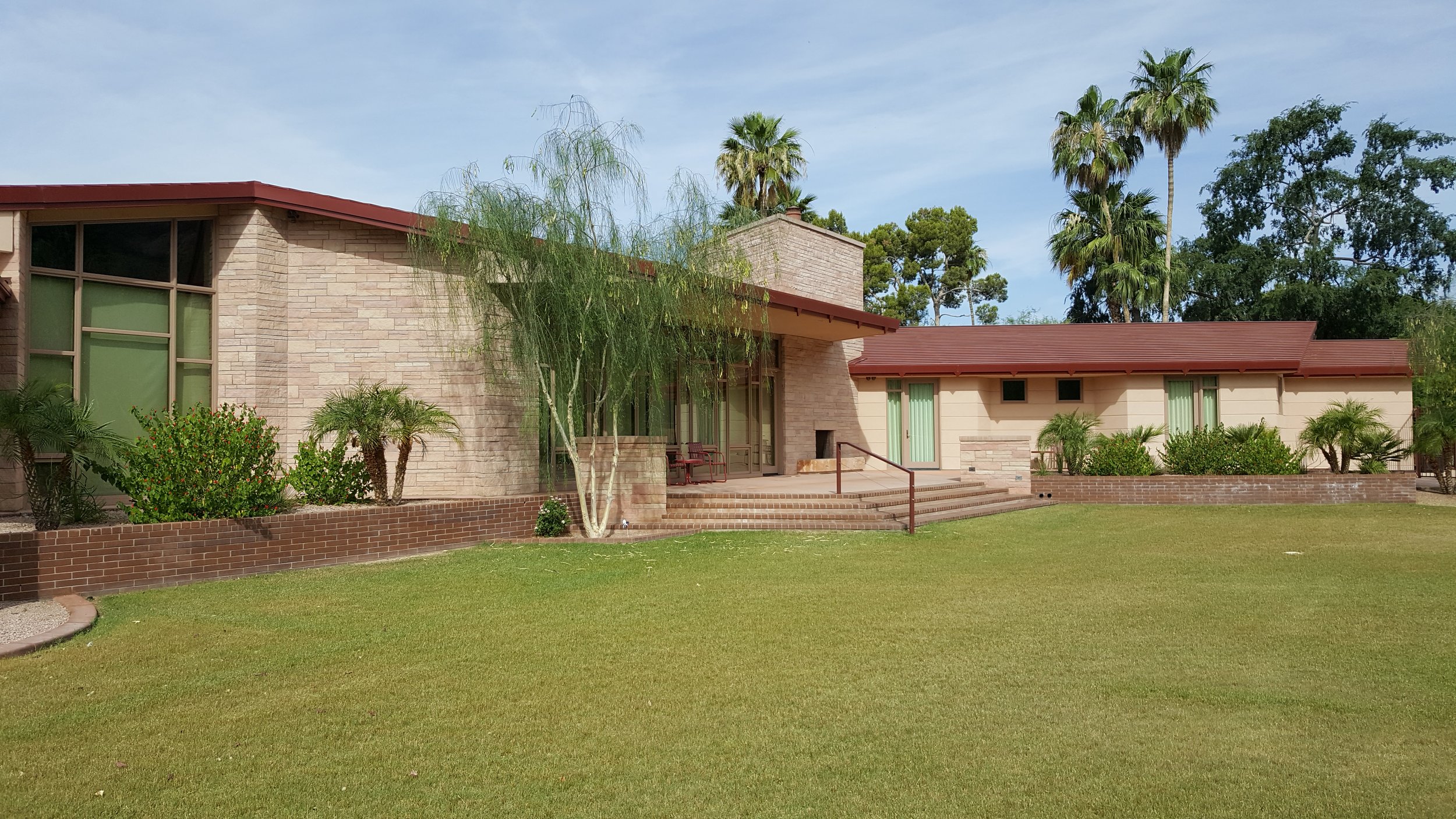 Modern house with stone masonry façade, pitched roof, and large windows surrounded by lush grass and palm trees.
